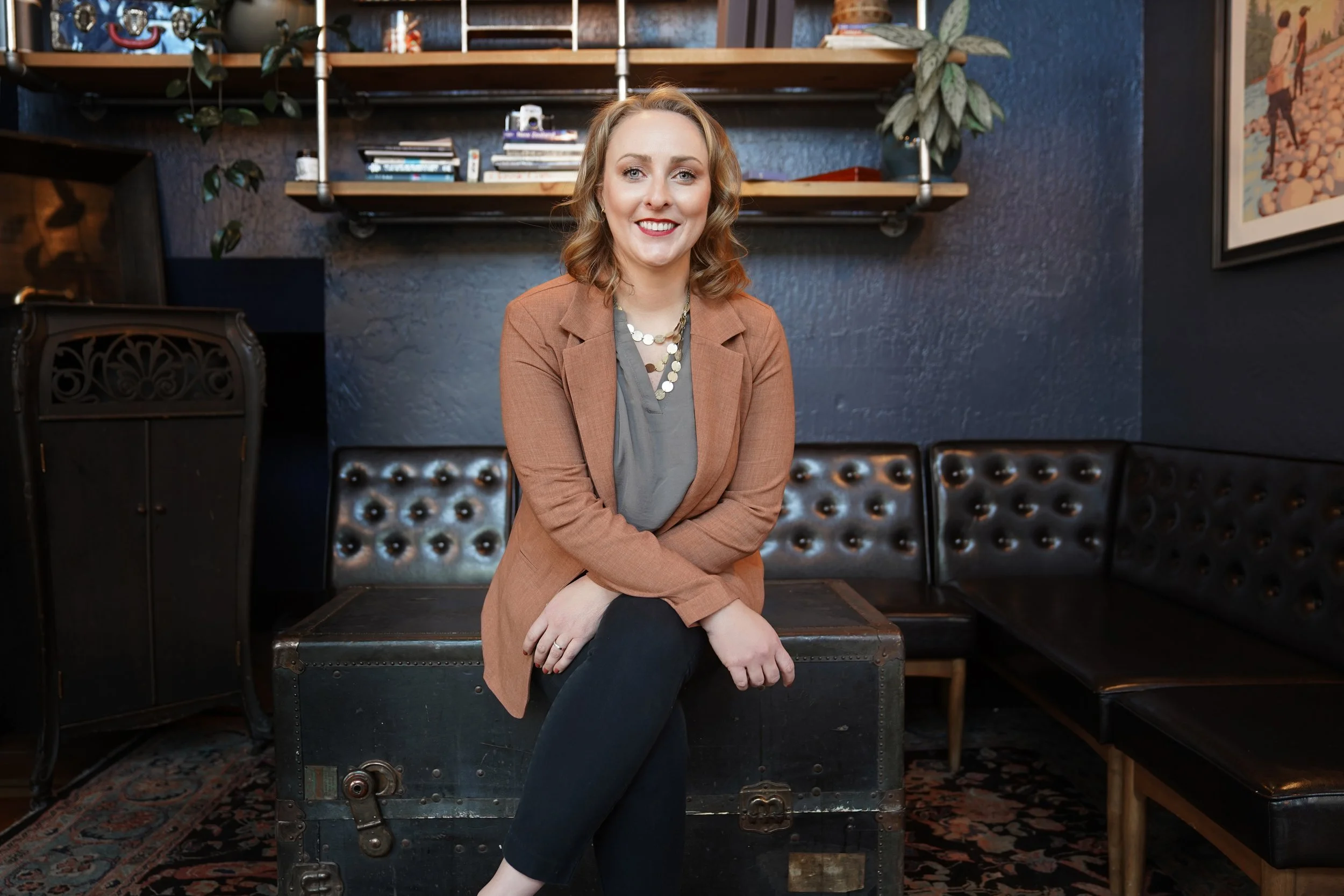 Diana Pullen, Redmond Oregon real estate broker in Central Oregonseated on a vintage trunk in a cozy room with dark blue walls, surrounded by books and plants.