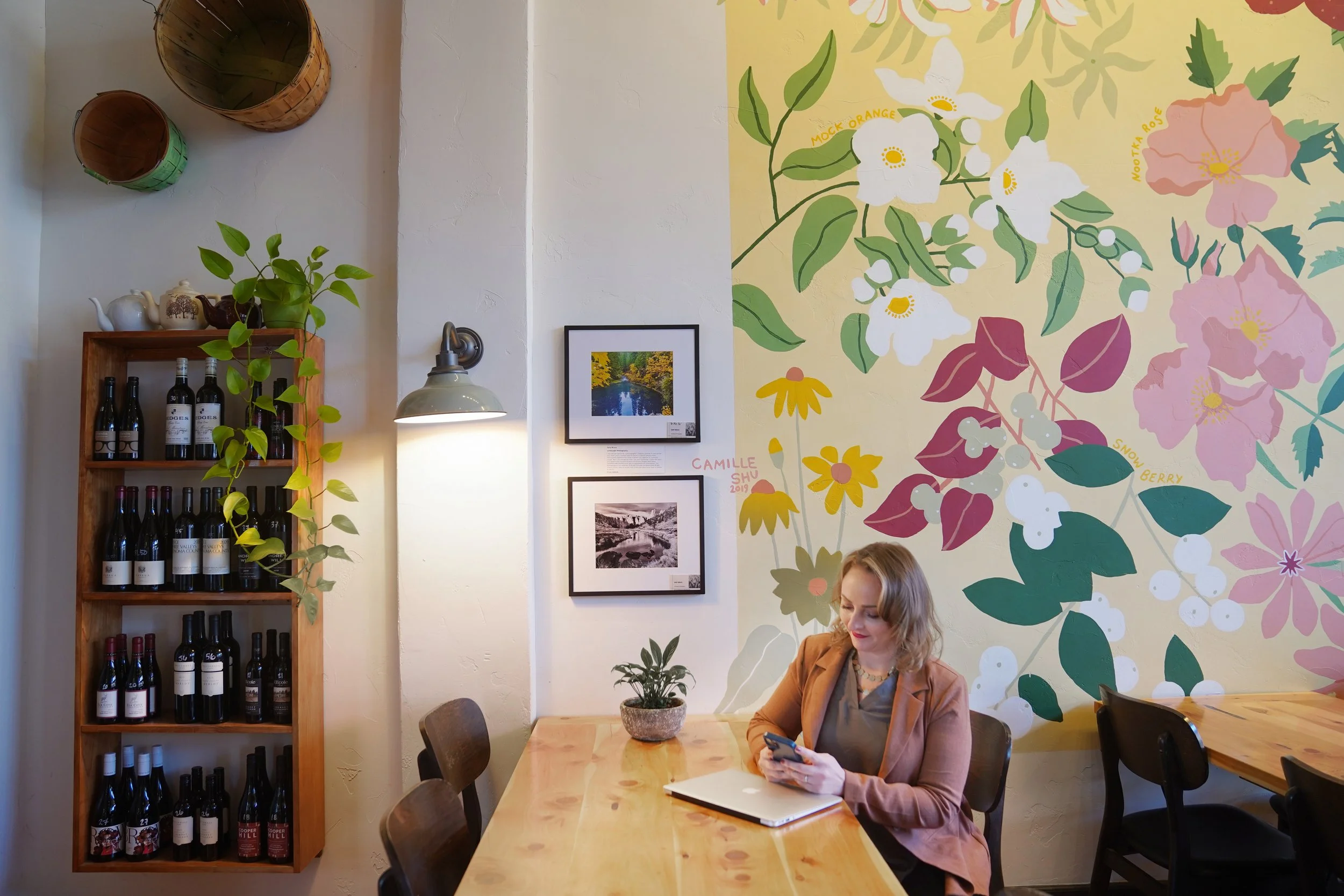 Diana Pullen real estate broker in Redmond Oregon sitting at a wooden table with a tablet and a notebook, in a cafe with colorful floral mural on the wall, a framed photograph, and a shelf with wine bottles and plants.