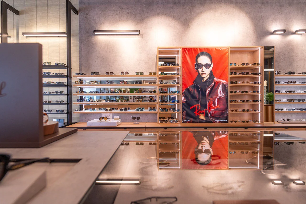 Nella vetrina di un negozio di occhiali si vedono molte mensole con diversi modelli di occhiali da sole e da vista, con un poster grande di una donna con occhiali Ray-Ban appeso al centro.