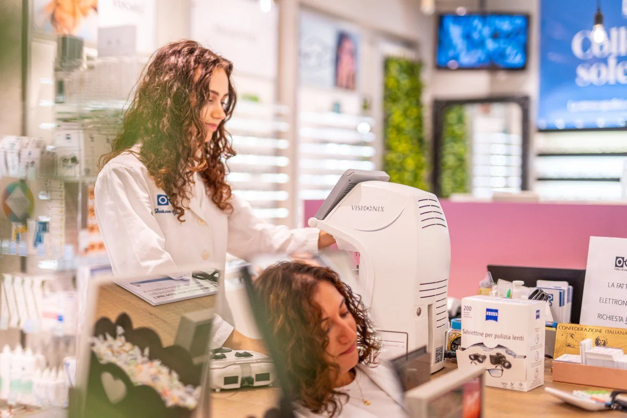 Una donna con i capelli ricci castani che si fa controllare gli occhi da un'ottica, mentre una cliente con capelli ricci si siede di fronte a una macchina ottica in un negozio.