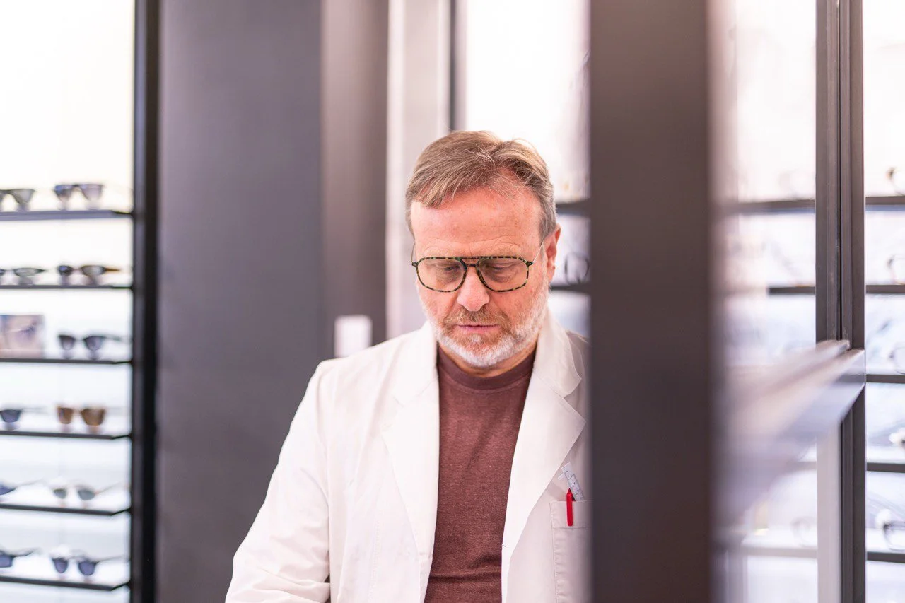 Uomo con capelli corti e barba, indossa occhiali retro e camice bianco in negozio di occhiali, osserva con attenzione una selezione di occhiali da vista o da sole.