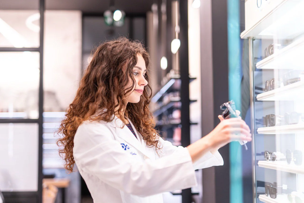 Una donna con capelli riccioli castani e occhiali da vista sta guardando un paio di occhiali presi da uno scaffale, indossando un camice bianco.