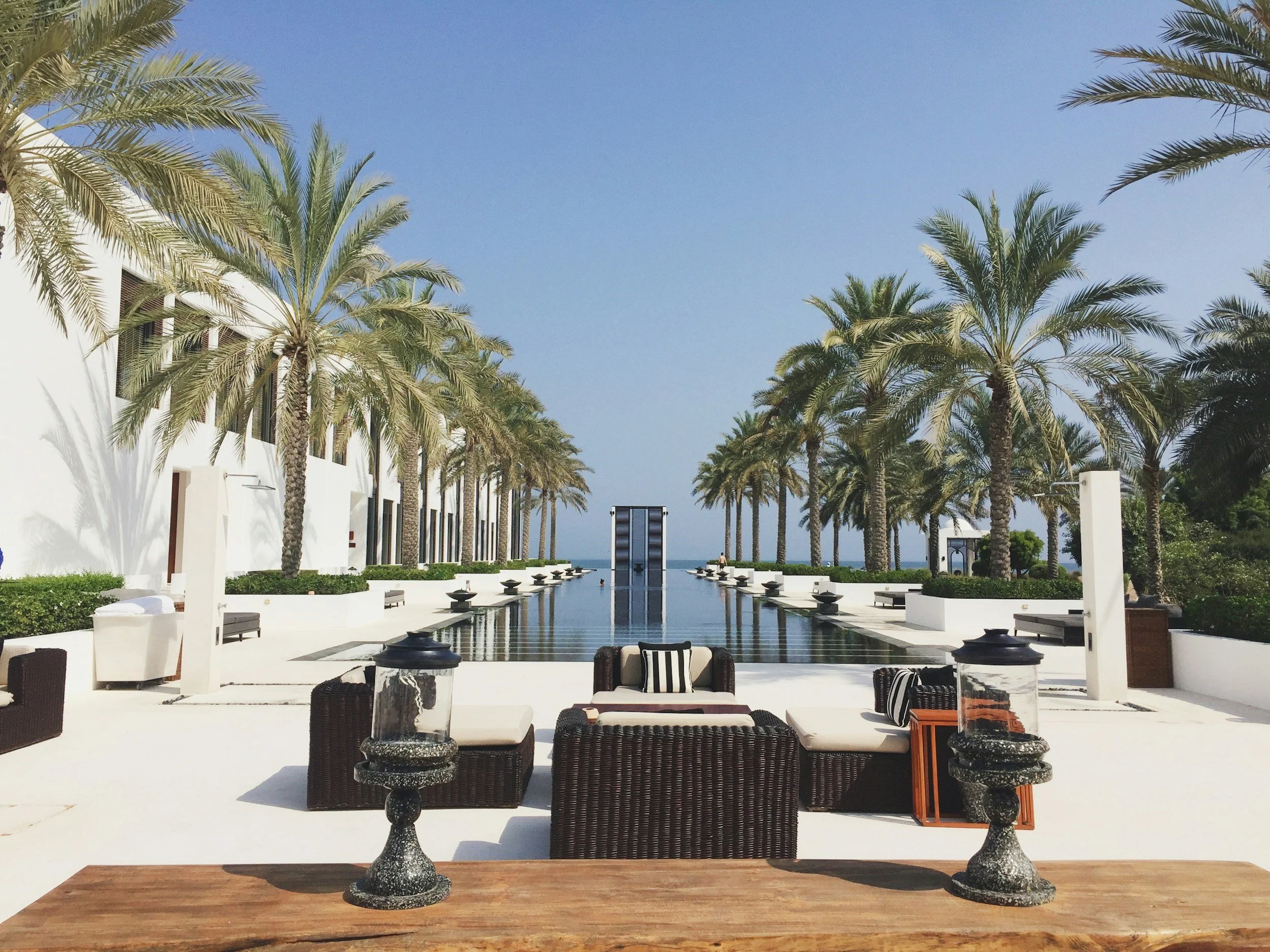 Luxury resort pool area surrounded by palm trees, with lounge chairs, umbrellas, and decorative lanterns, overlooking the ocean in the background.