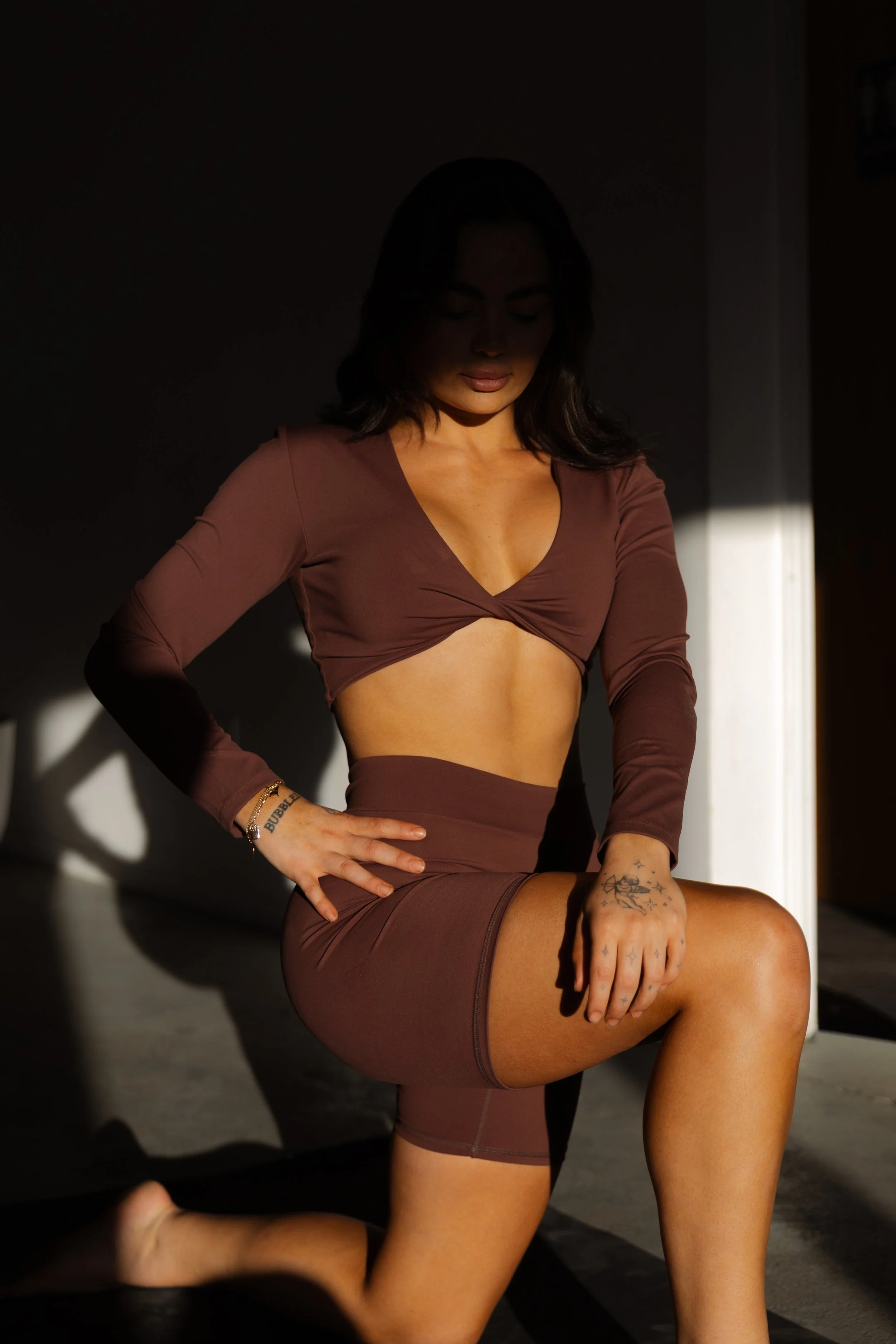 A woman in a brown crop top and matching shorts kneeling on the ground in sunlight with shadows in the background.