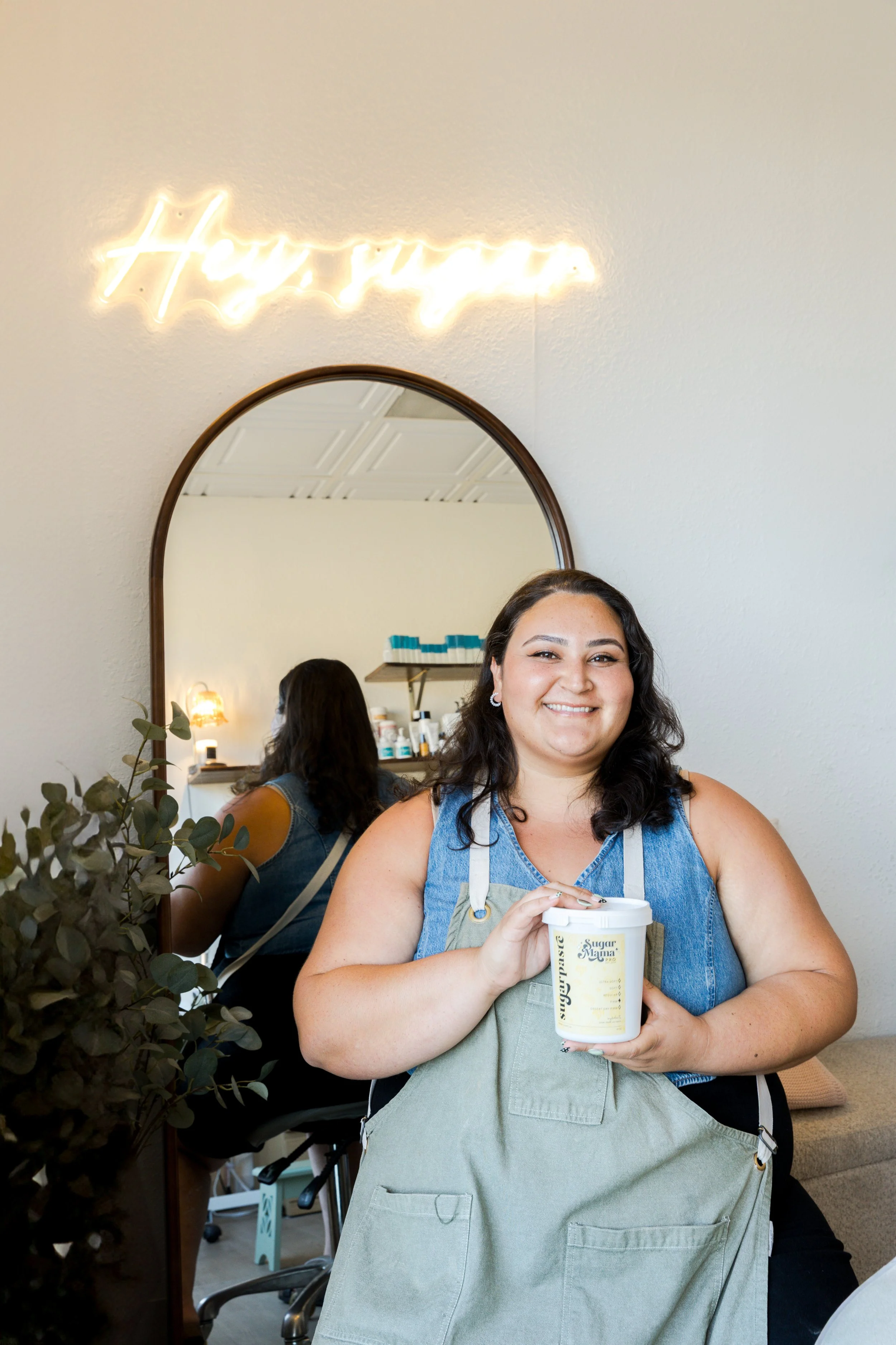 A smiling woman with dark, wavy hair wearing a denim sleeveless top and a beige apron, holding a container of sugar scrub, standing in a well-lit room with a large mirror, neon sign in the background, and plants.