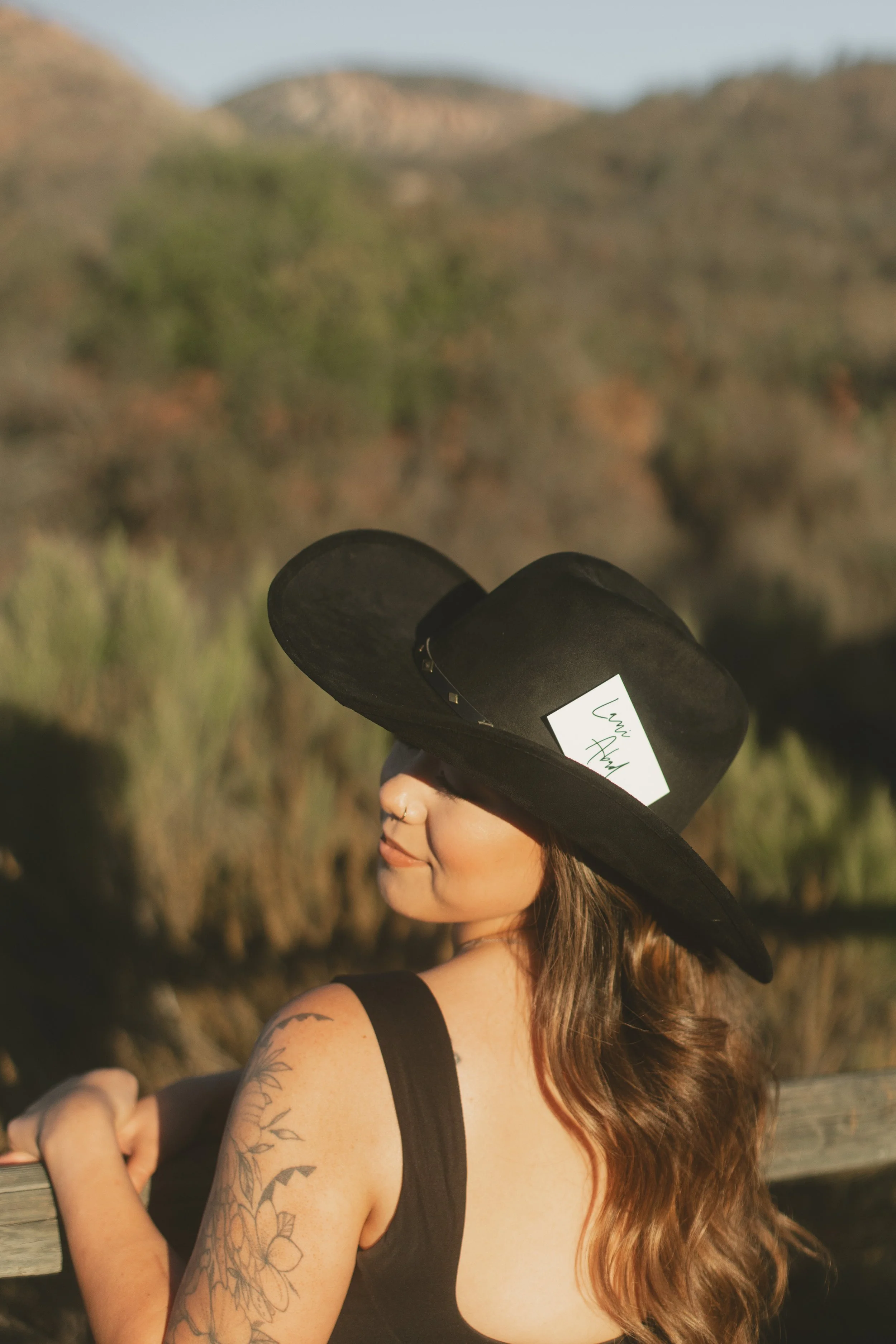 A woman with long brown hair, tattoos on her shoulder, and a septum piercing, wearing a large black hat with a white tag, standing outdoors with hills and desert vegetation in the background.