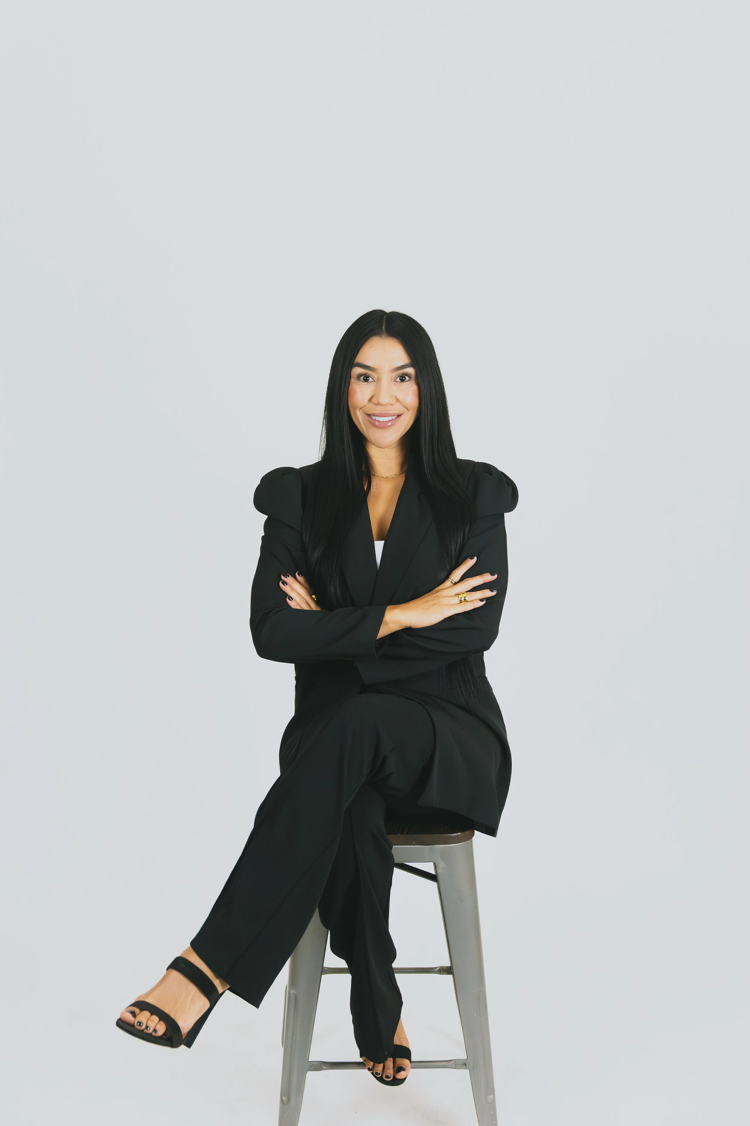 A woman in a black suit sitting on a high stool with crossed arms, smiling, against a plain light gray background.