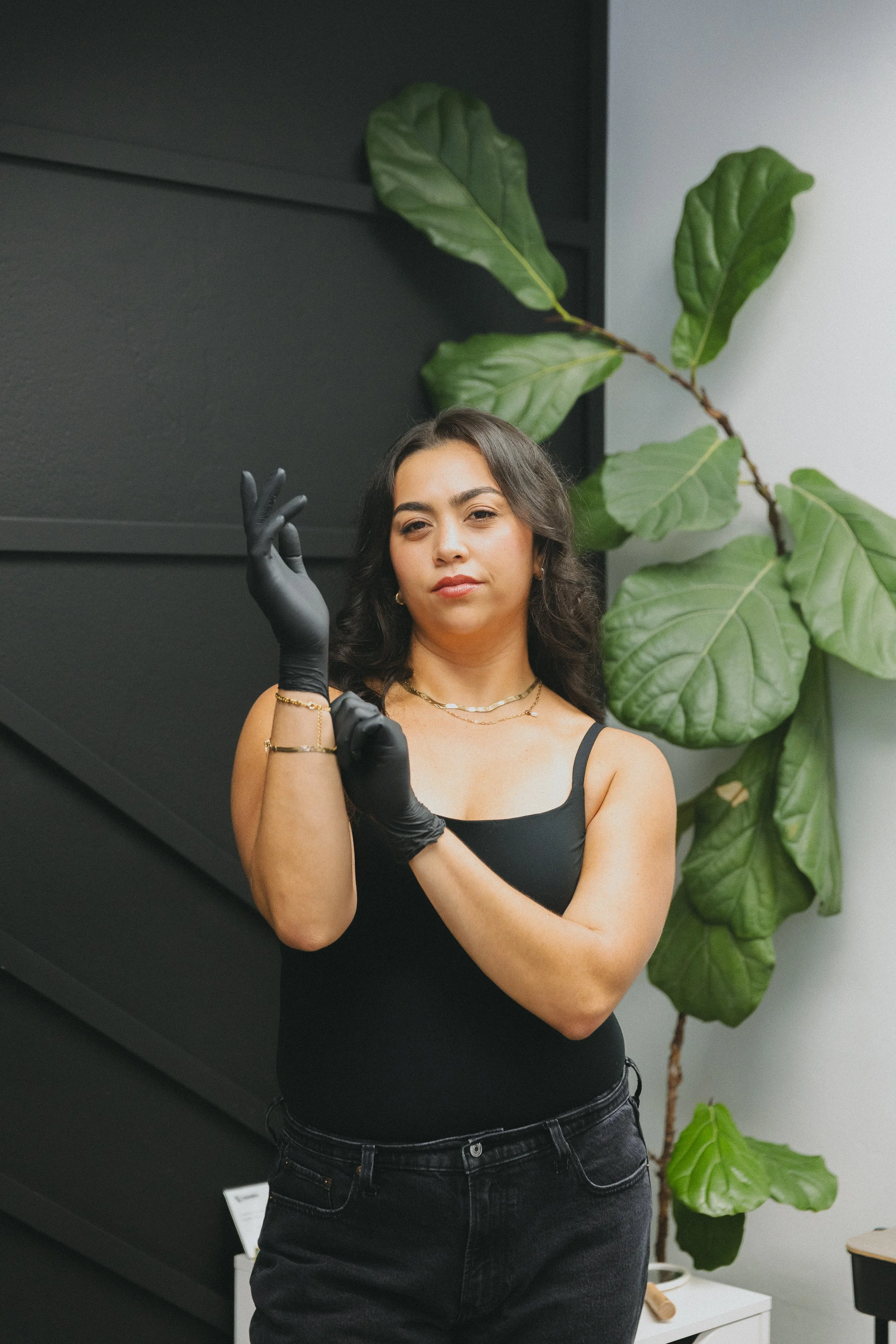 A woman wearing a black tank top, black jeans, and black gloves, standing in front of a black wall and large green plant, looking at the camera with a composed expression.