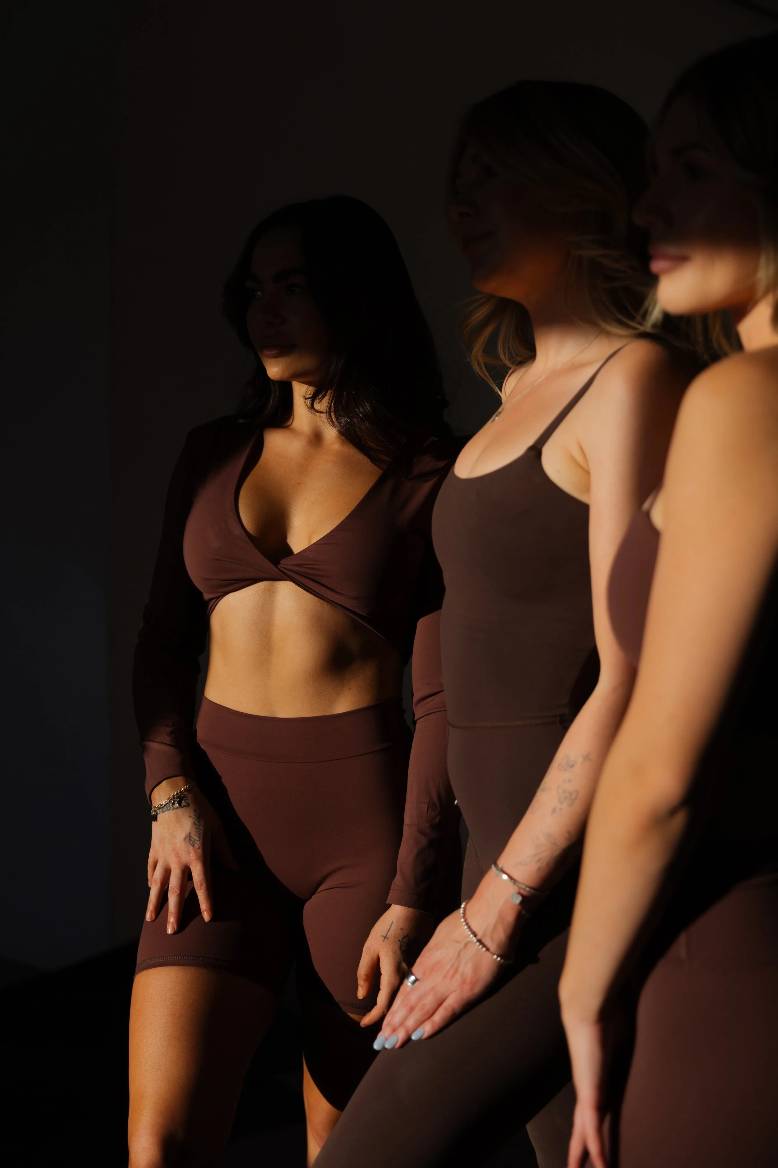 Three women standing side by side in dark lighting, wearing brown and black athletic outfits.