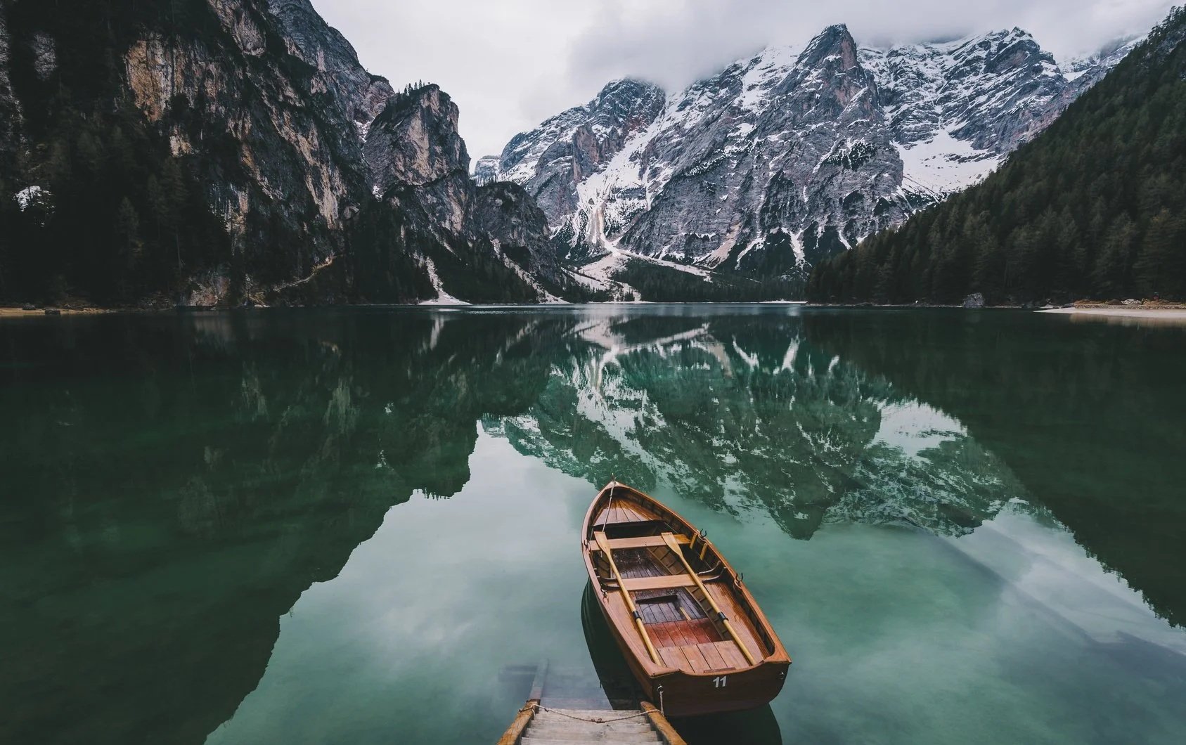 A tranquil lake surrounded by snow-capped mountains and dense evergreen forest, with a wooden boat tied to a dock in the foreground, and a mirror-like reflection of the mountains on the water.