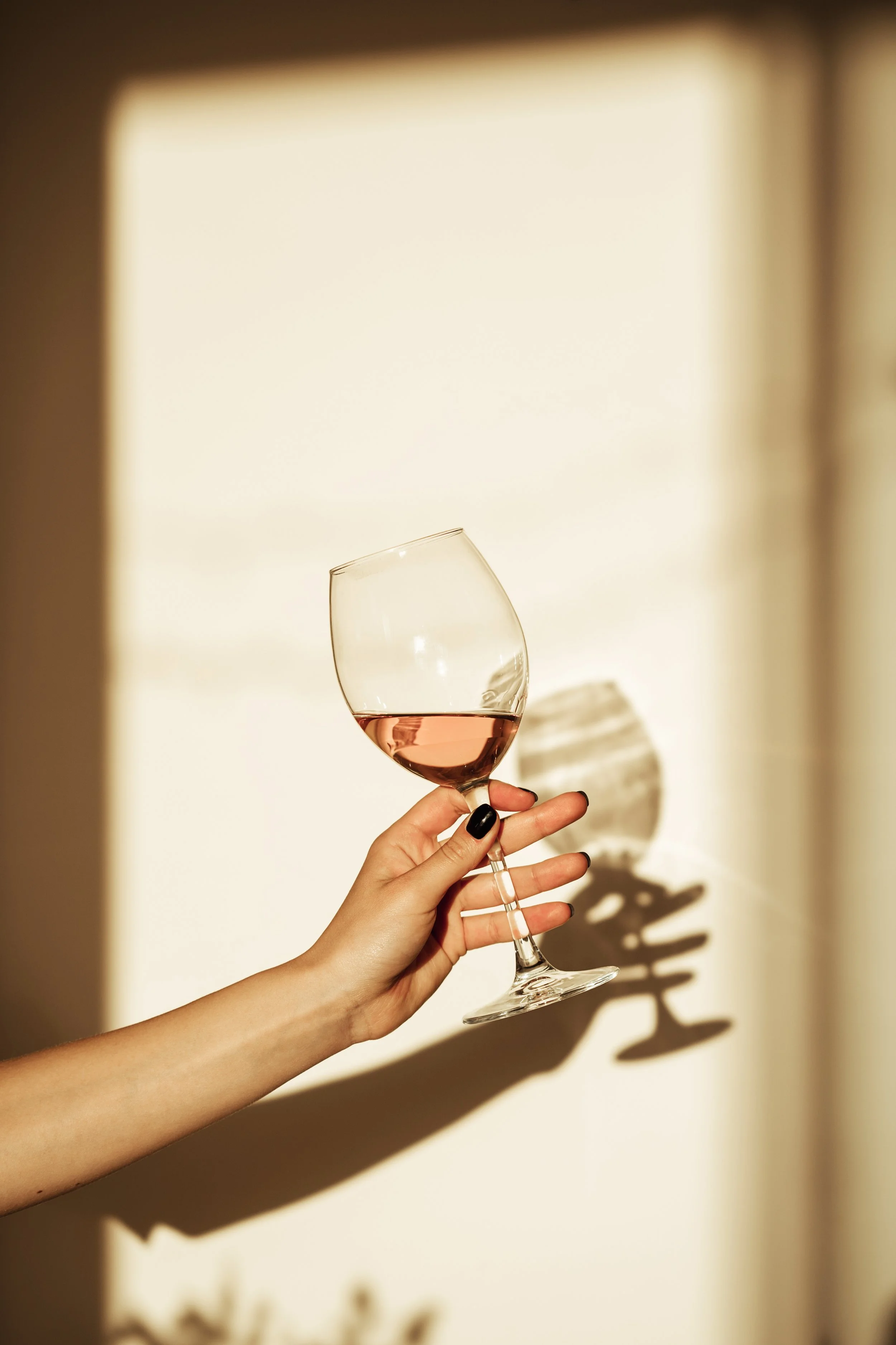 A hand holding a glass of rosé wine against a white background with the shadow of the glass cast on the wall.