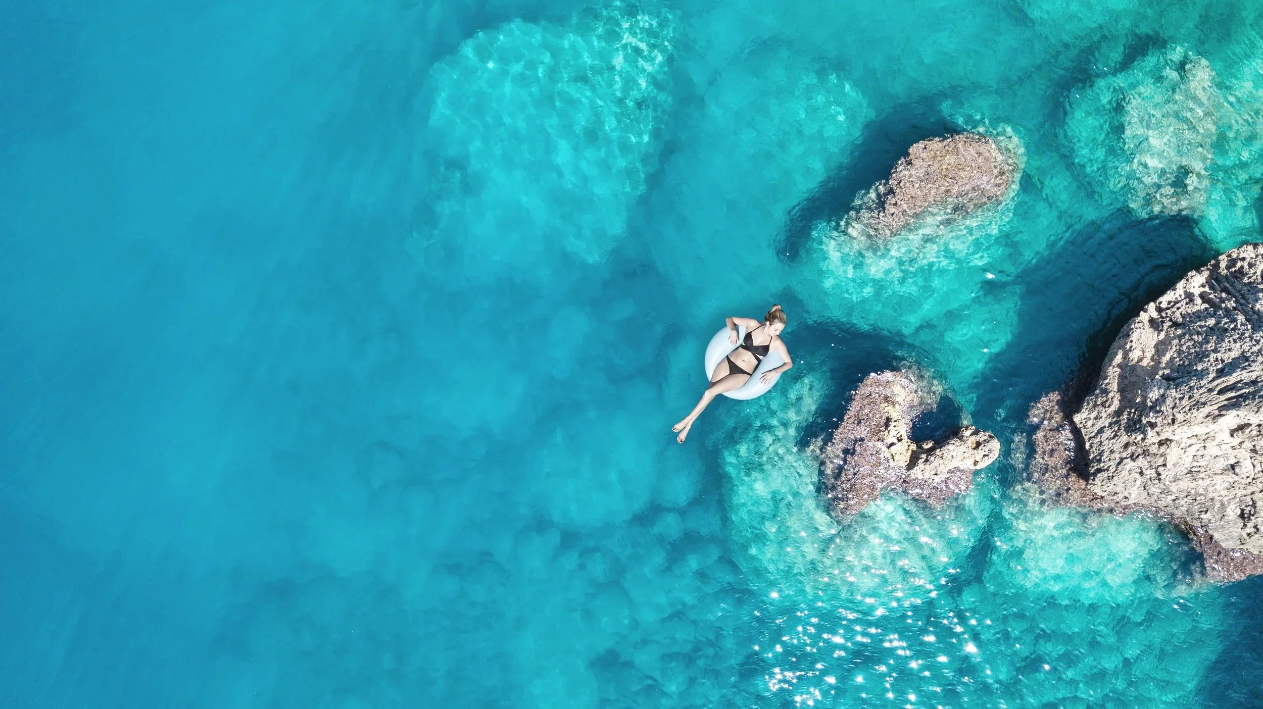 A woman in a black swimsuit relaxing on an inflatable ring in clear blue ocean water near rocky formations.