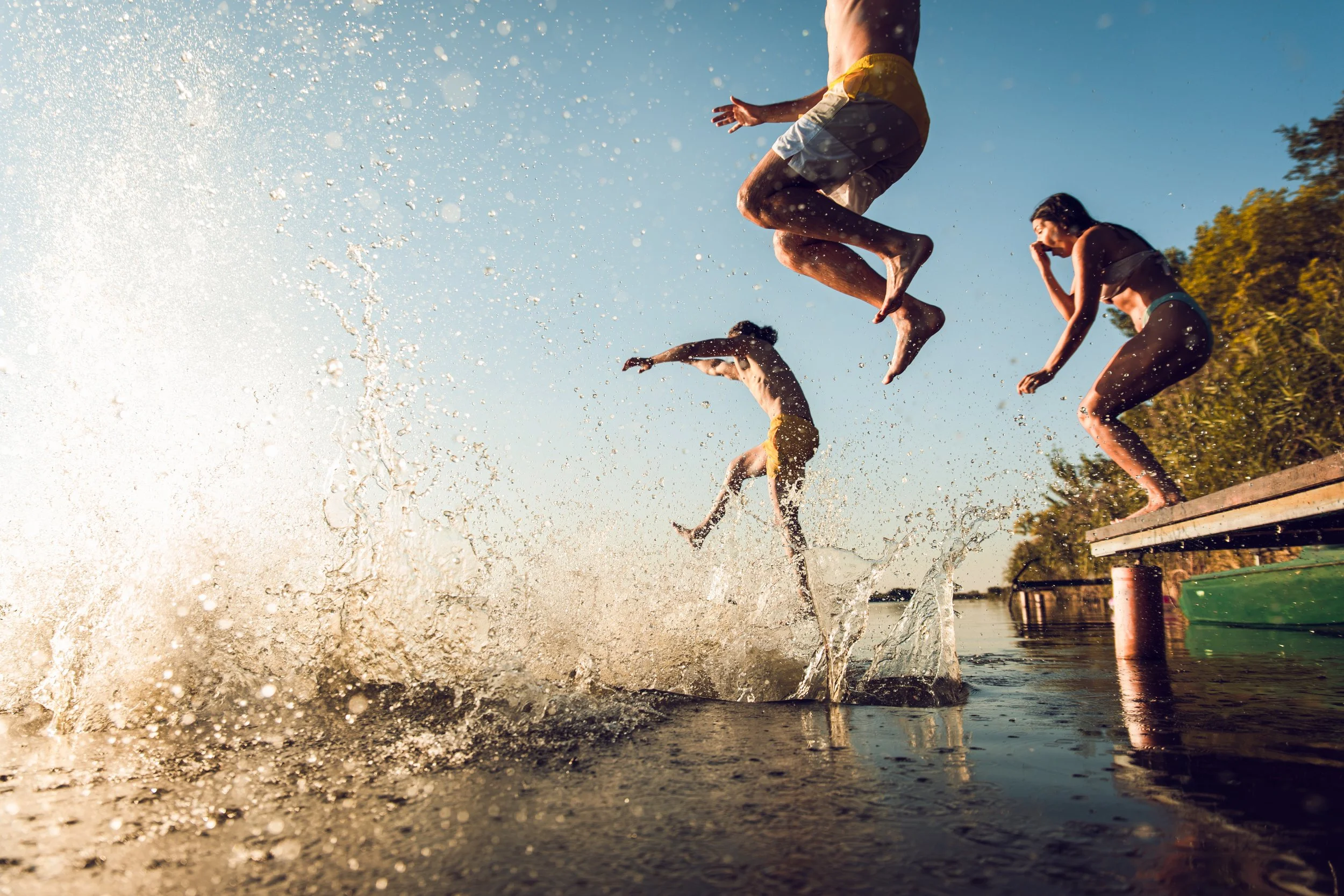 Three children jumping into water with splashes, under a blue sky in a park or lake setting.