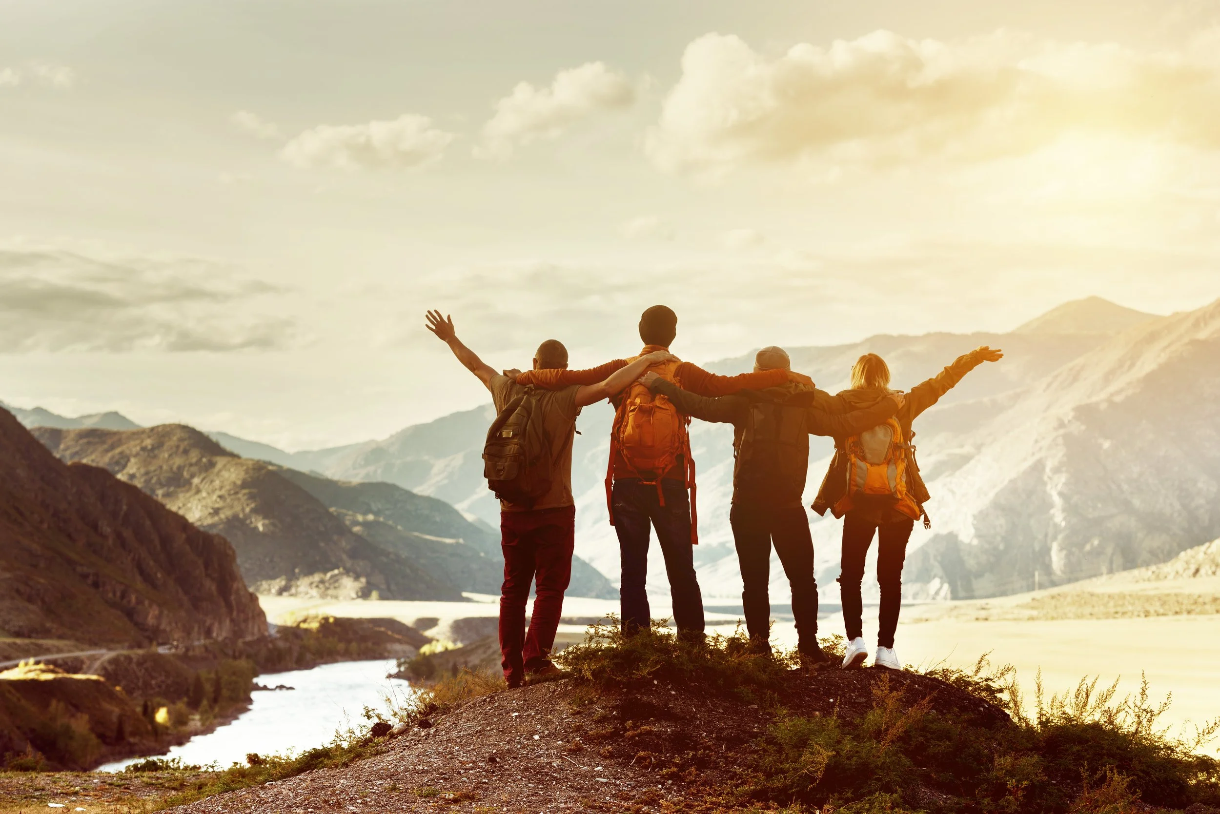 Group of five friends with backpacks standing on a hilltop overlooking mountains and a river, celebrating with arms raised during sunrise.