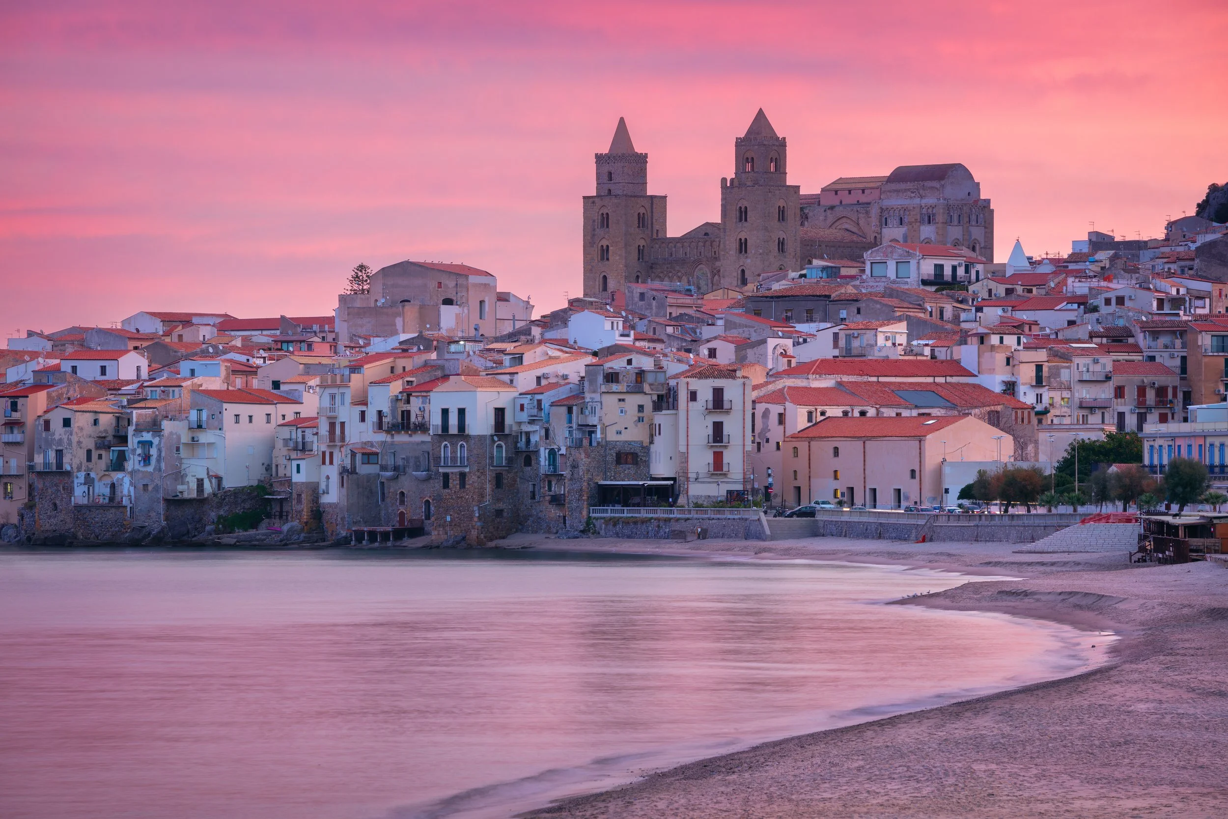A scenic view of a coastal town at sunset with a historic church or cathedral with twin towers on a hill overlooking the buildings below. The sky is pink and purple, and the water in the foreground reflects the colors of the sky.