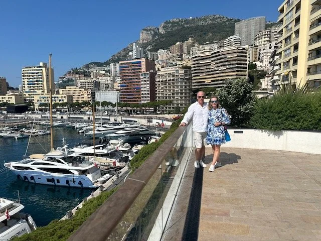 A couple standing on a waterfront terrace with yachts docked below, and tall buildings with mountainous terrain in the background.
