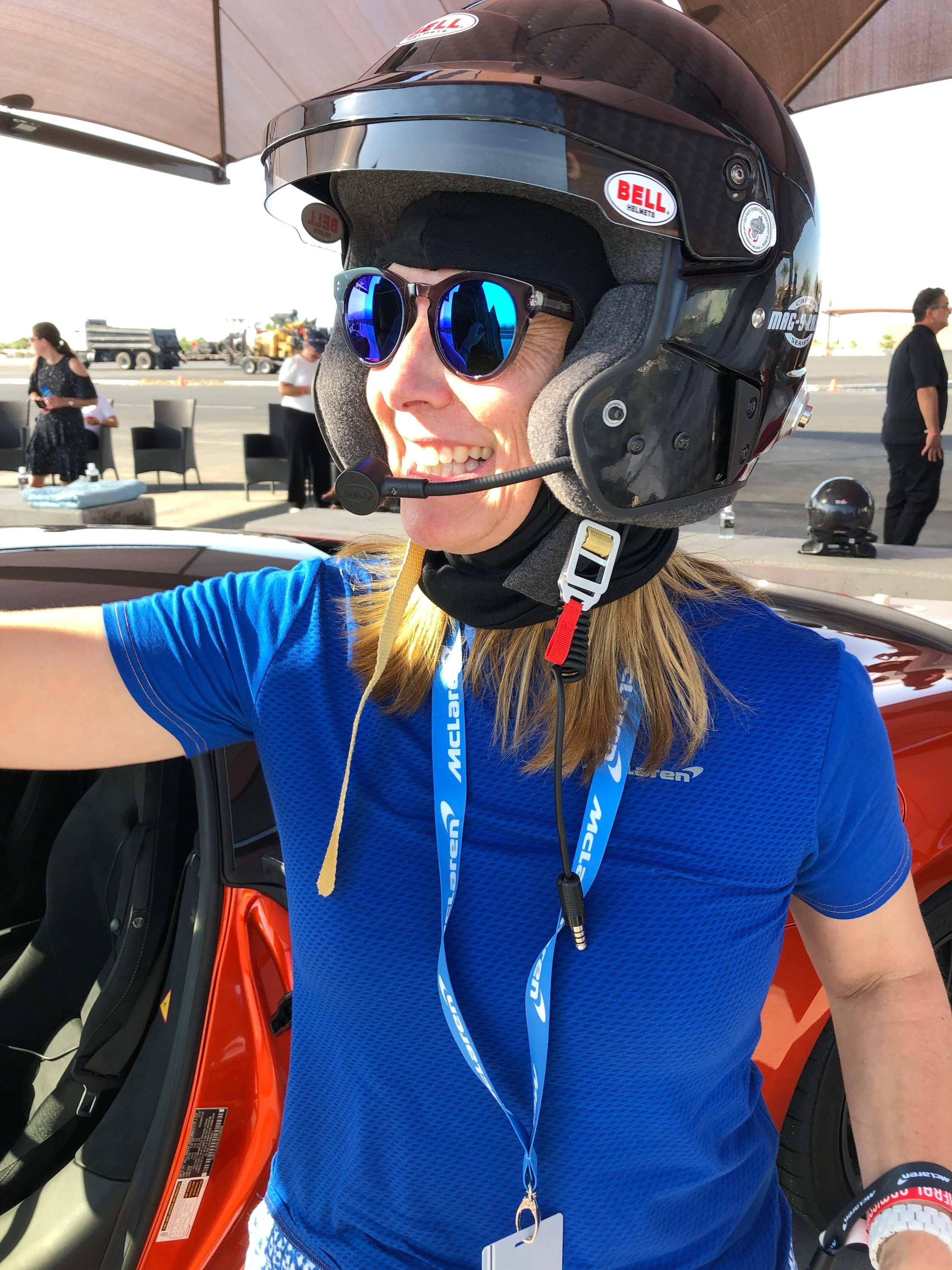 Person smiling, wearing a racing helmet and sunglasses, standing next to a race car at a racing event.