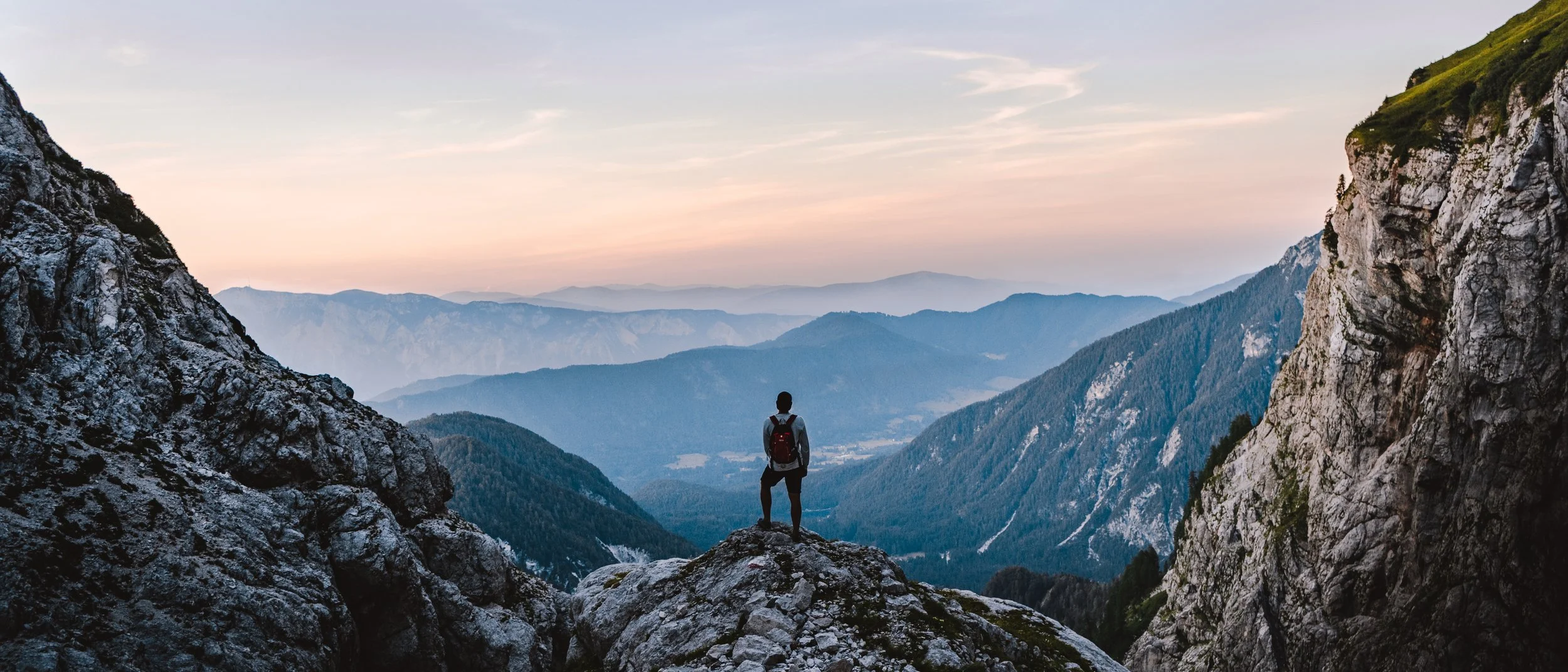 A person standing on a rocky ledge overlooking a valley with mountain ranges in the distance during sunset.