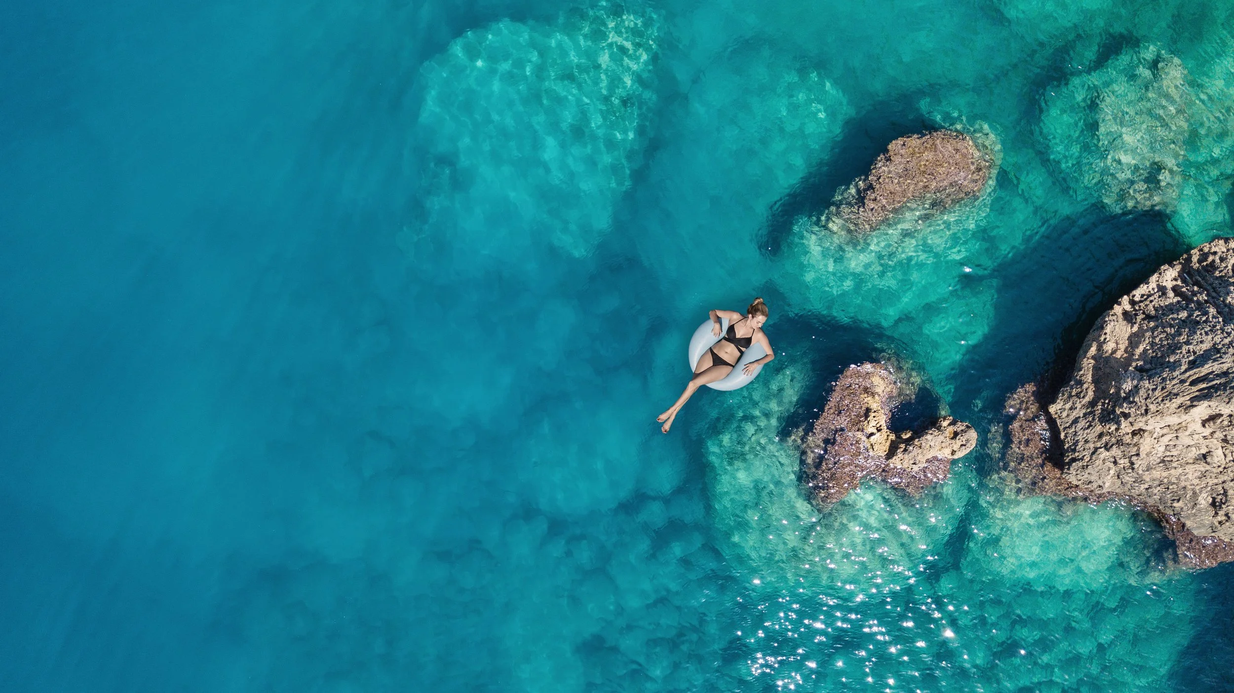A woman in a black bikini floating on a white pool float in clear turquoise water near rocky formations.