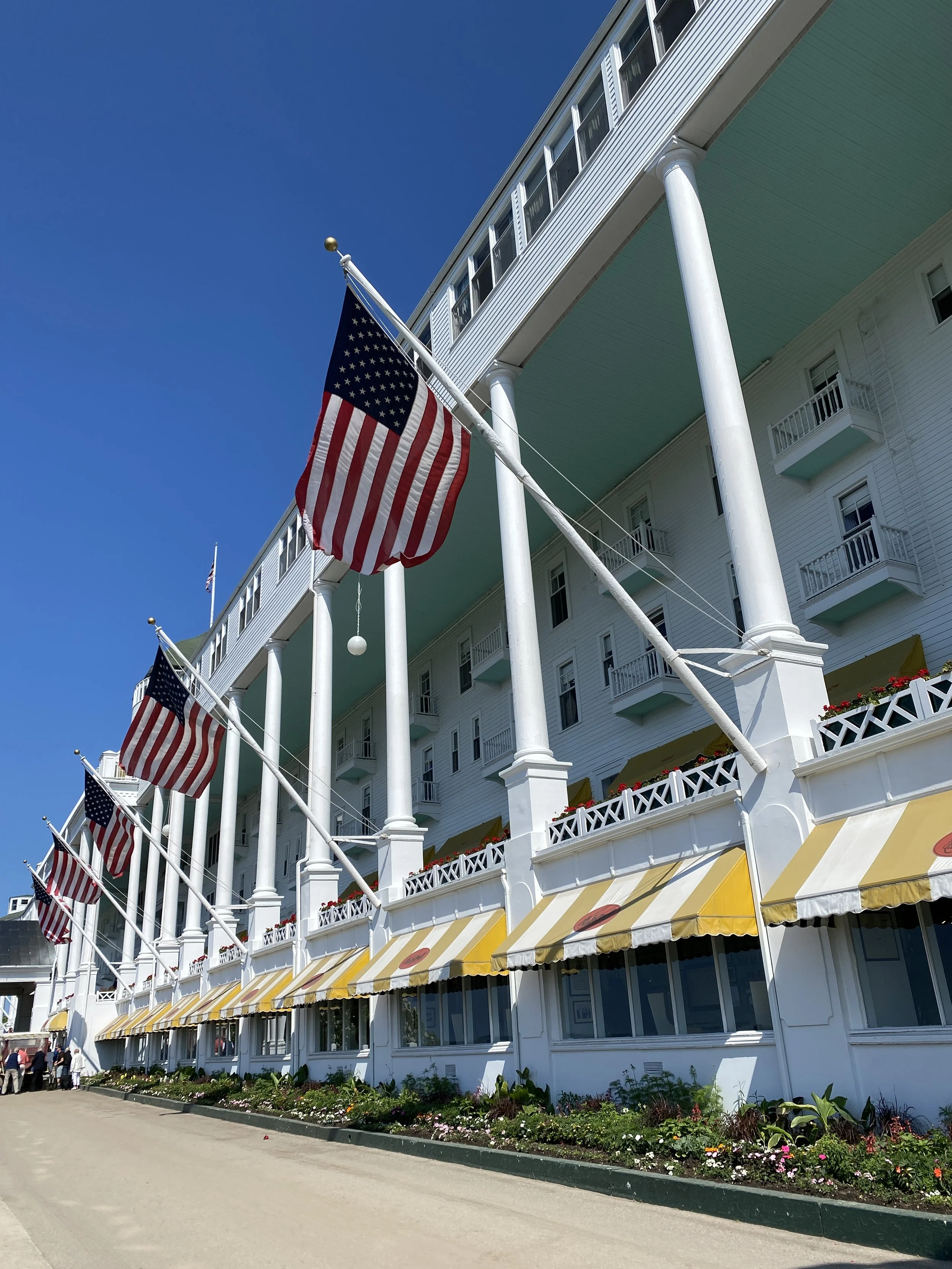 Exterior of a large white building with multiple balconies, decorated with American flags on flagpoles and yellow striped awnings, set against a clear blue sky.