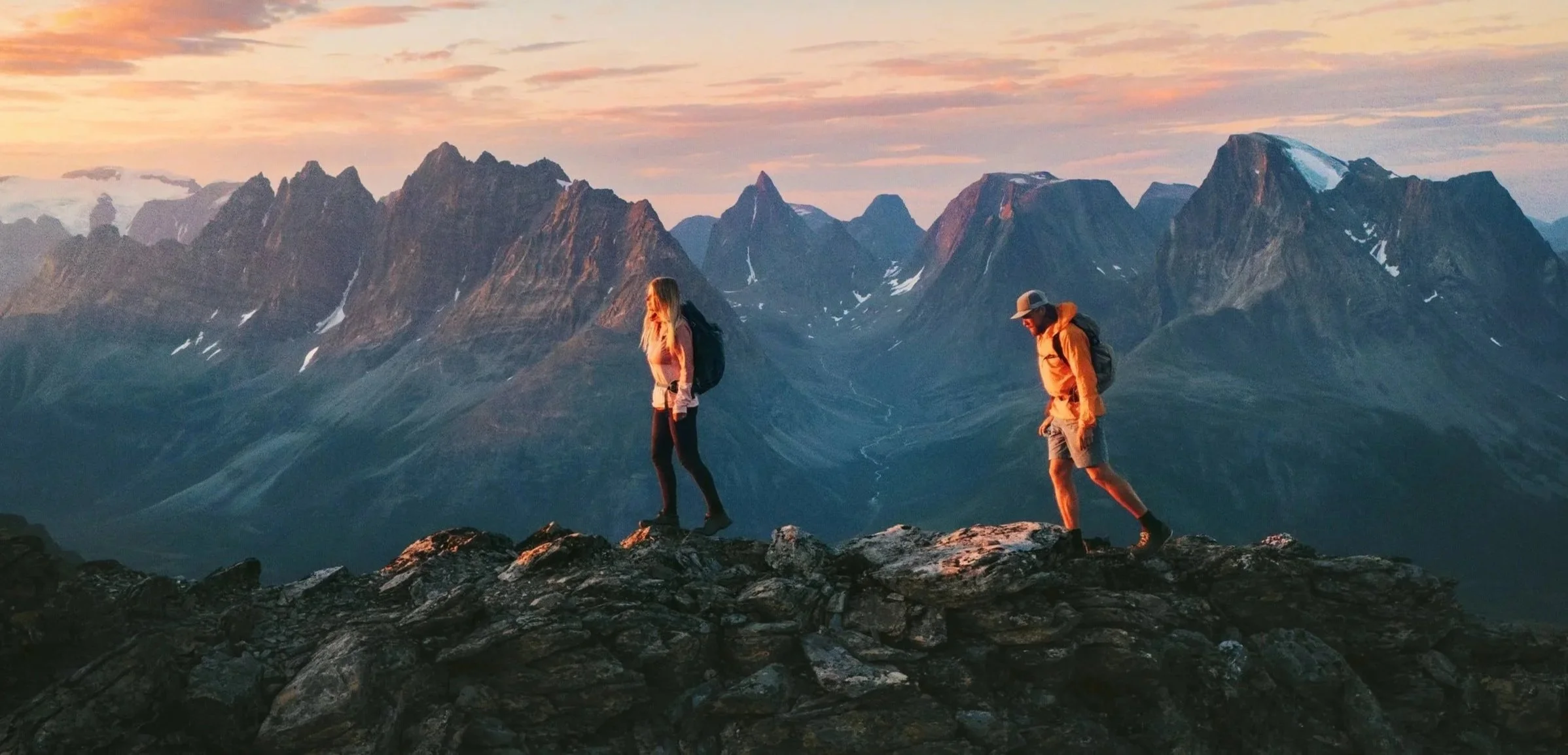 Two hikers walking on rocky terrain with a mountain range and sunset in the background.