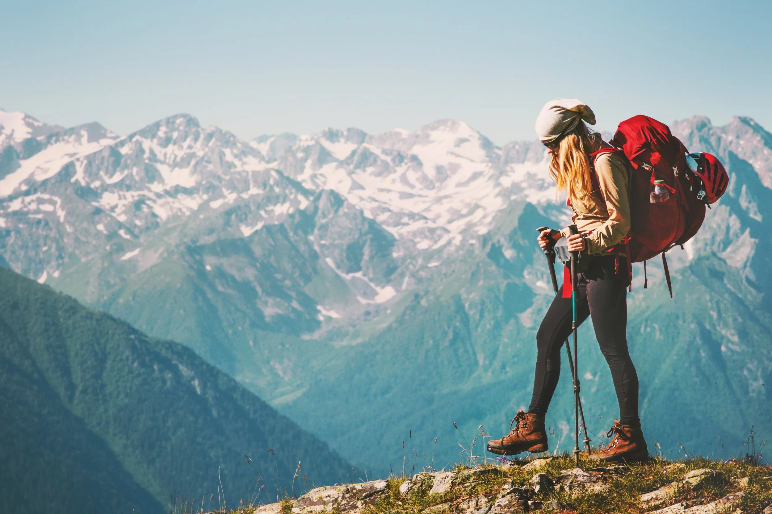 A hiker with long hair wearing a gray beanie, sunglasses, a beige jacket, black pants, and brown hiking boots standing on a rocky ledge with snow-capped mountains in the background.