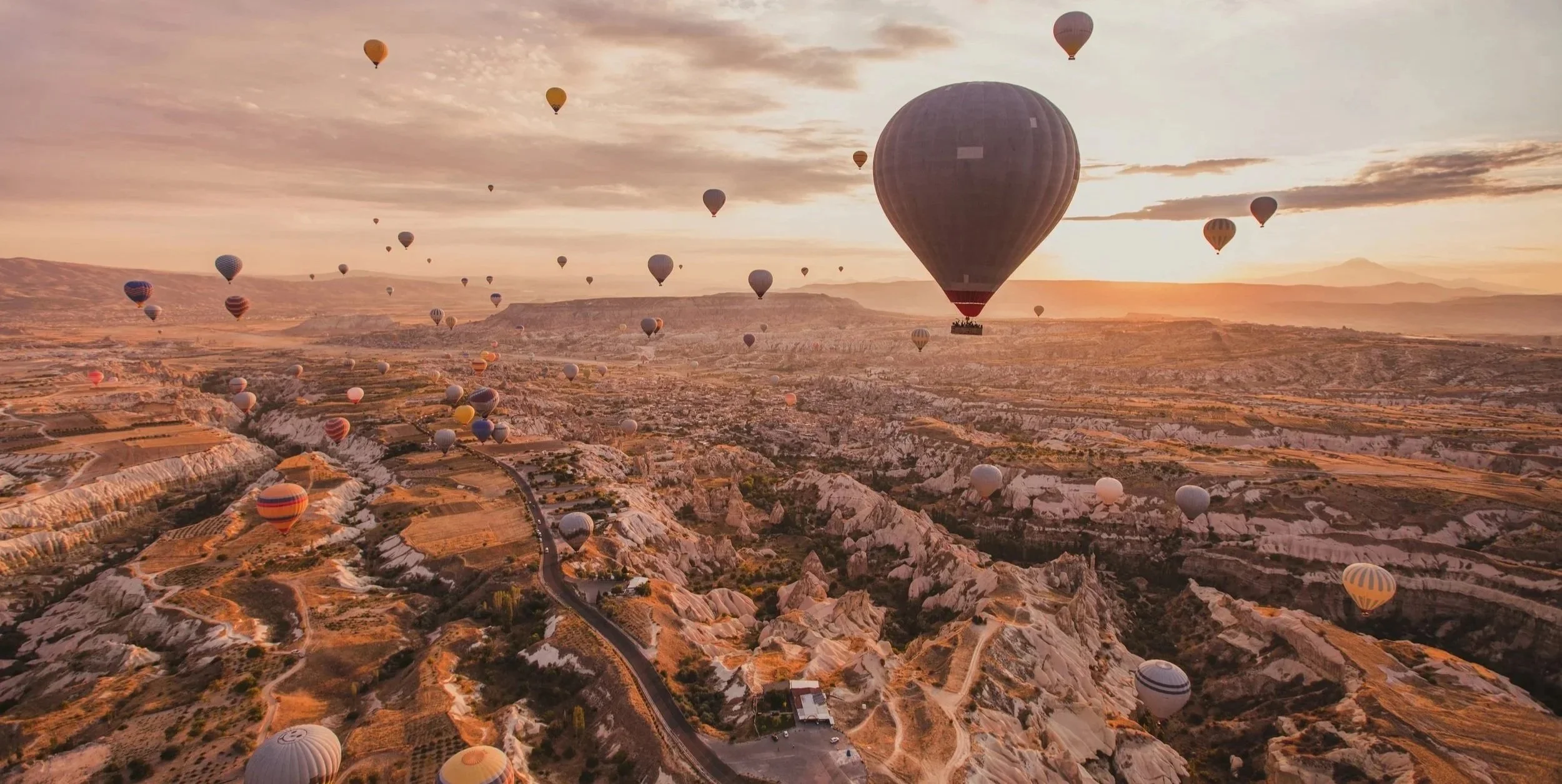 Aerial view of numerous hot air balloons floating over a rugged landscape during sunrise or sunset, with hills and valleys in the background.