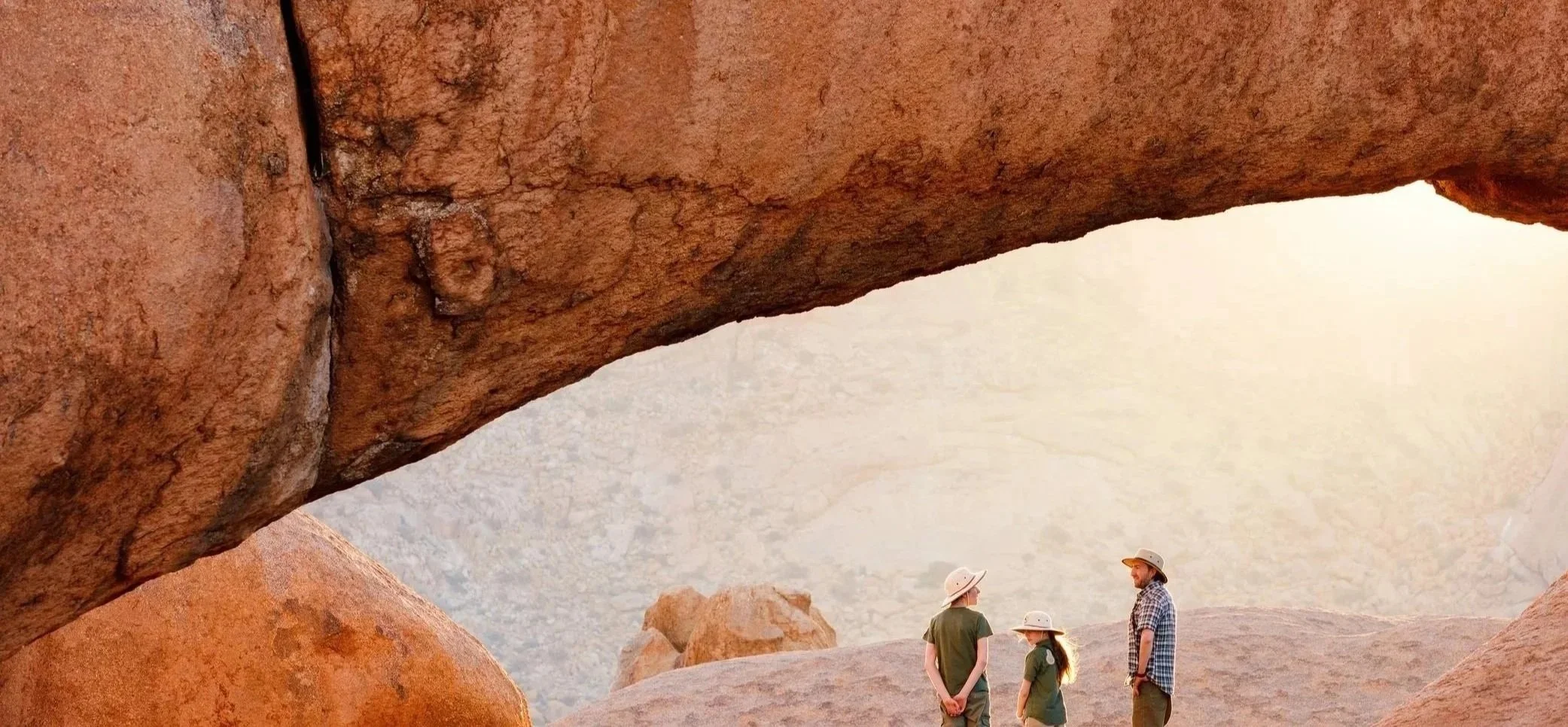 Three people wearing hats standing and talking beneath a large, curved rock formation in a desert landscape with reddish rocks and mountains in the background.
