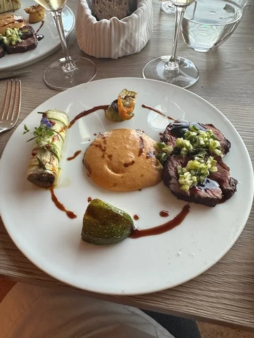 A white plate with a rolled appetizer garnished with herbs, a small roasted vegetable, a scoop of creamy sauce, and grilled meat topped with chopped vegetables, with sauce drizzled on the plate. Two wine glasses and a water glass are on the table behind it.