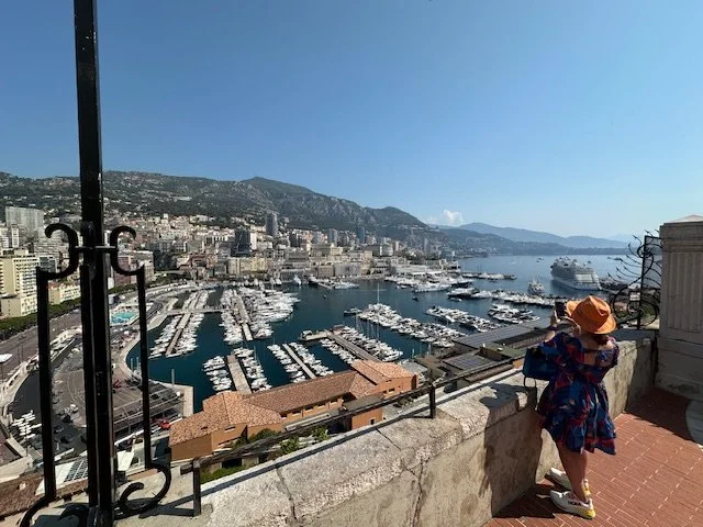Child wearing a hat and colorful dress looking over a harbor filled with yachts and boats, with a city and mountains in the background.