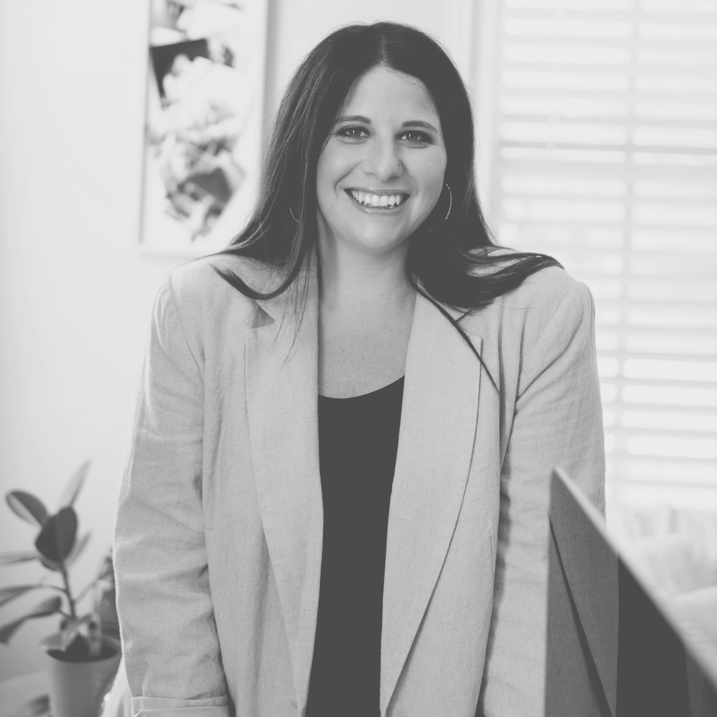 Black and white photo of a Blake Grun in an office setting, wearing a light blazer and hoop earrings, with a plant and window blinds in the background.