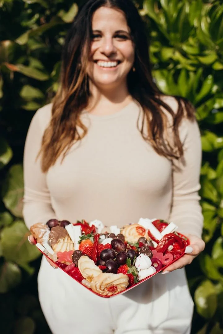 Woman smiling and holding a plate of assorted desserts including strawberries, grapes, cookies, and whipped cream in front of green foliage.