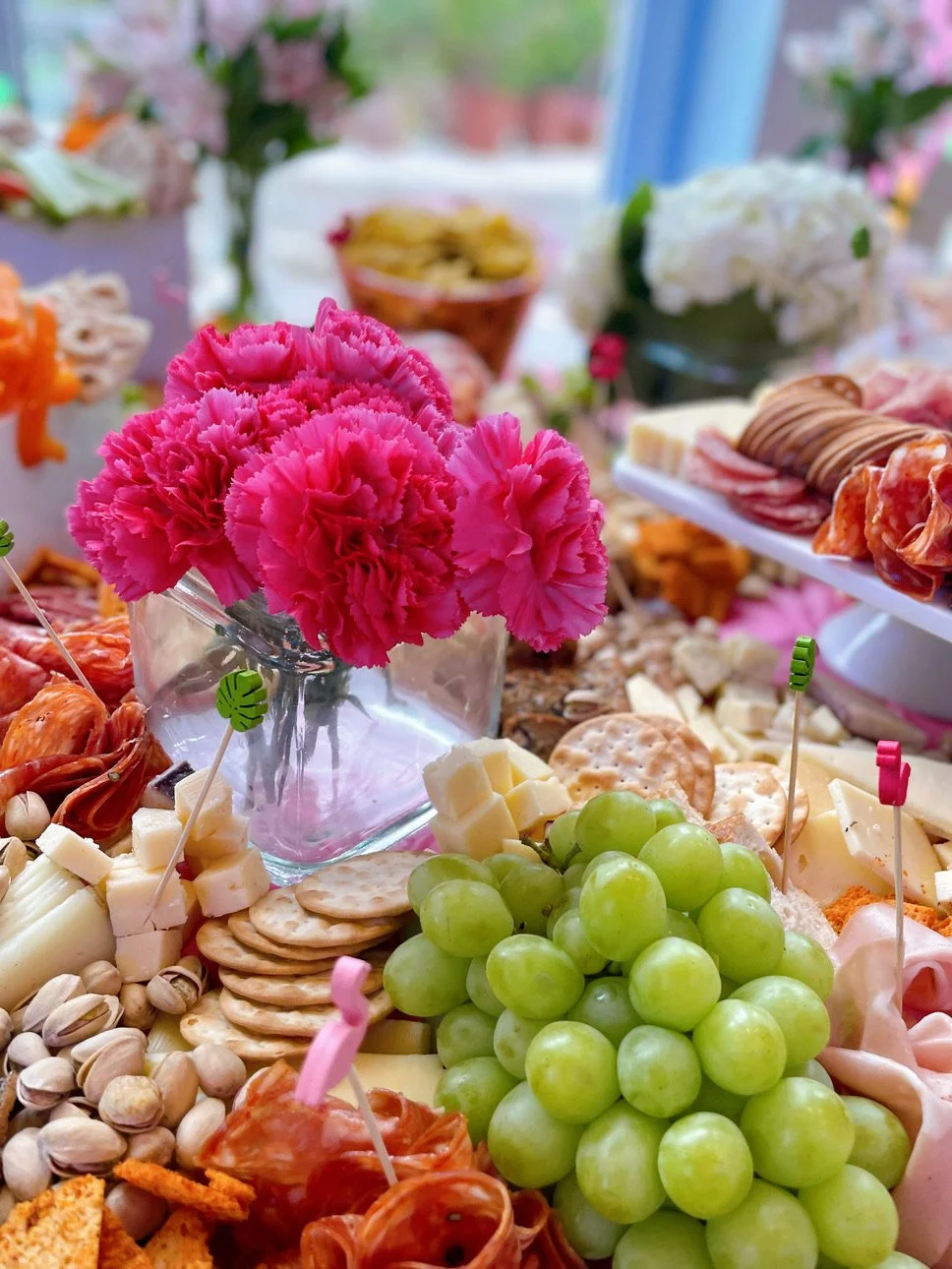 Colorful charcuterie board with green grapes, pink flowers in a jar, cheese cubes, nuts, crackers, and cured meats