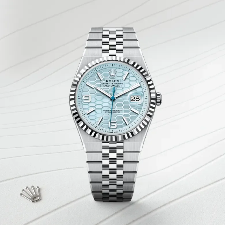 A Rolex Oyster Perpetual Land Dweller wristwatch with a light blue patterned dial and a two-tone metal bracelet, laid on a white textured surface with a small Rolex crown logo nearby.
