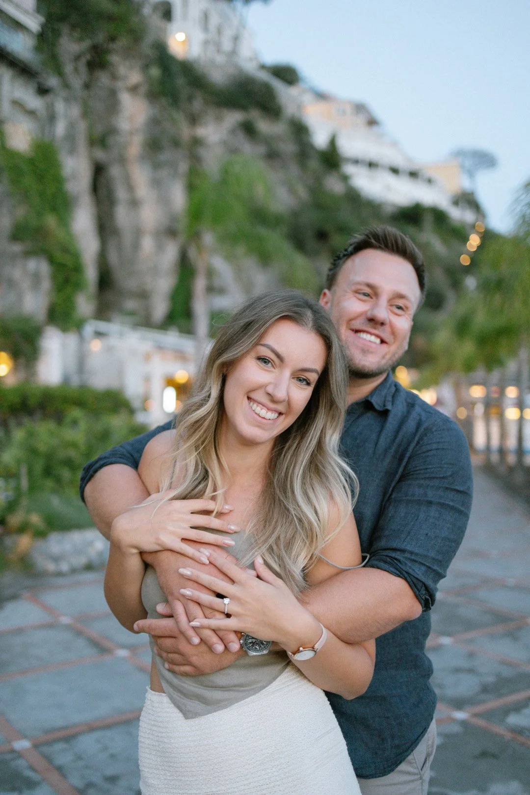 A smiling couple embracing outdoors in front of a hillside with houses, trees, and lights during the evening.
