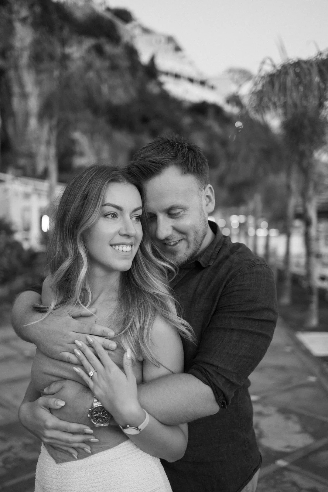 A happy couple embracing outdoors, with scenic mountains and trees in the background, in black and white.