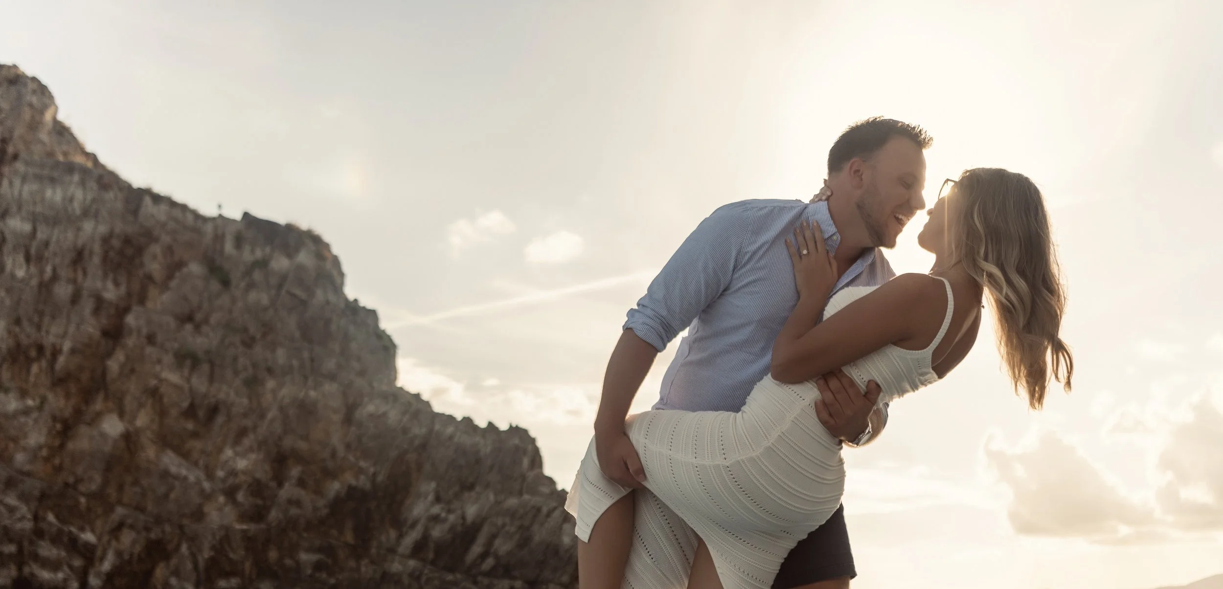 A man holding a woman in a dress on a rocky beach at sunset, both smiling and leaning close to each other.