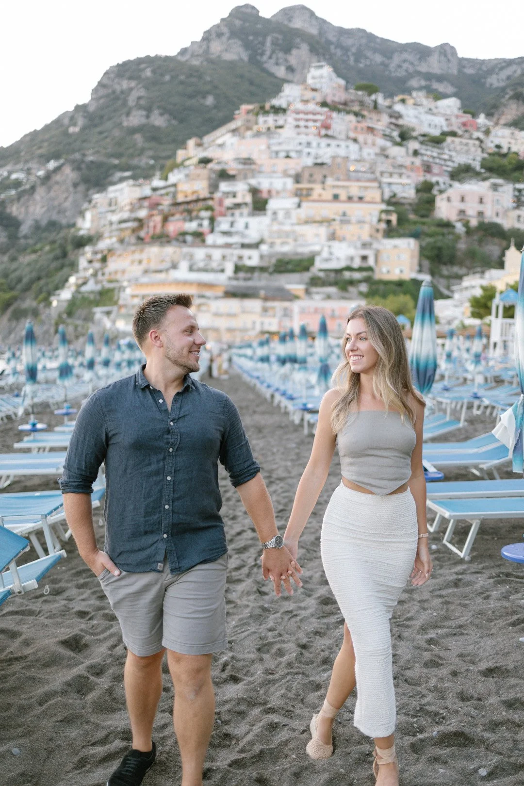 A young couple holding hands and walking on a beach with rows of blue lounge chairs and umbrellas. In the background is a hillside with colorful buildings.