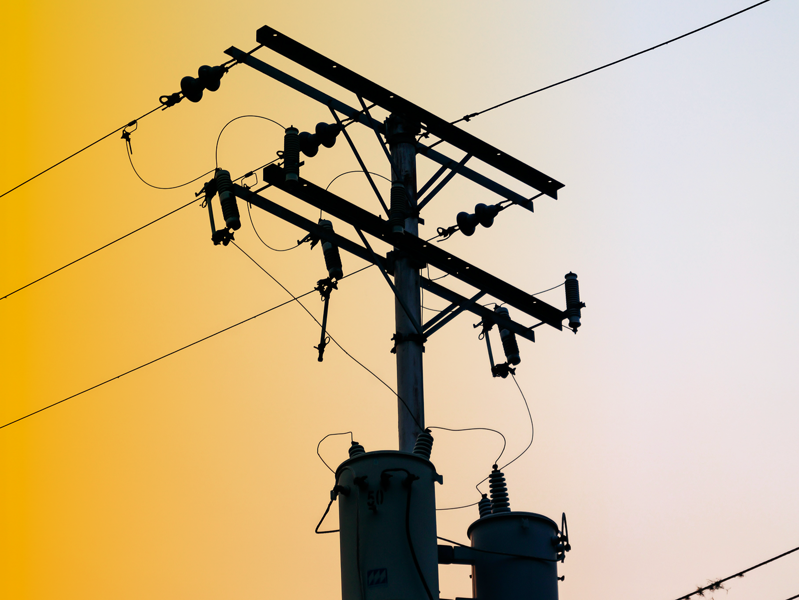 Silhouette of a power line pole with electrical insulators and wires against a sky with a yellow-orange gradient on the left fading to white on the right.