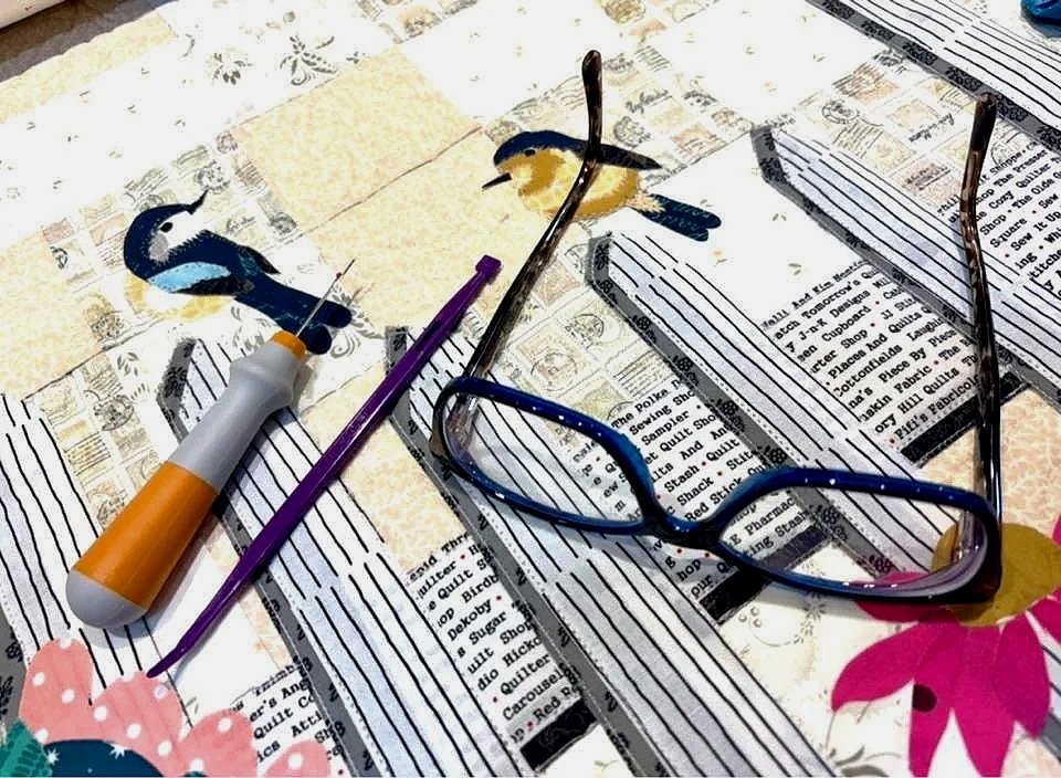 A pair of glasses, a syringe, a purple tool, and two bird illustrations on printed newspaper pages with colorful floral and bird-themed designs.