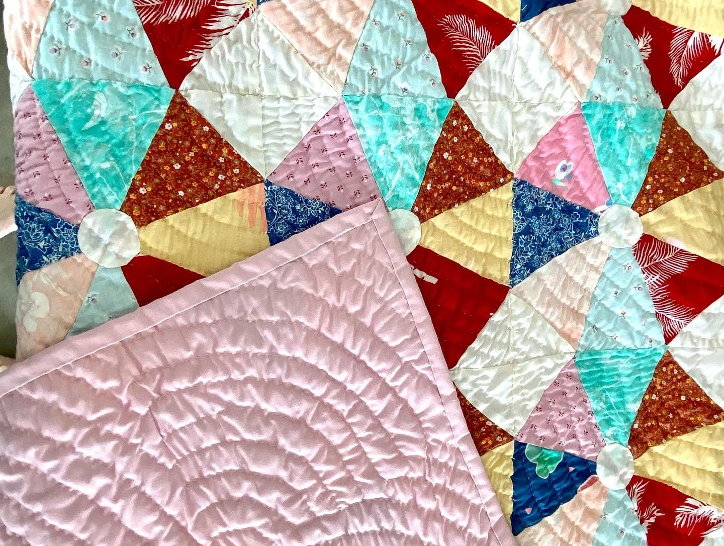 Two quilts with colorful geometric patterns and floral designs, one with a pink quilted fabric draped over a quilt featuring bright triangles and circles.