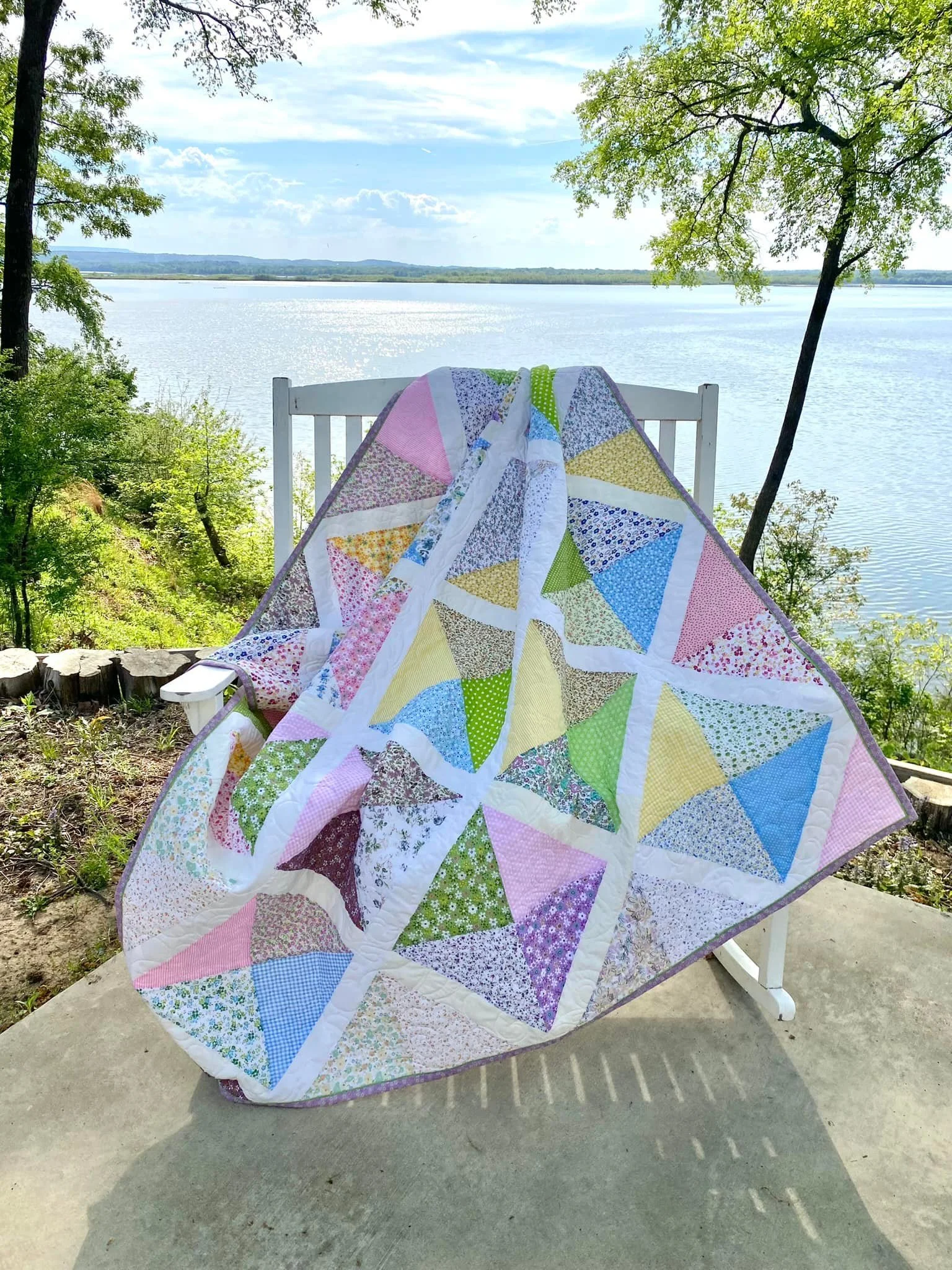 A colorful patchwork quilt with floral and striped patterns draped over a white rocking chair on a concrete patio near a river, with trees and a partly cloudy sky in the background.