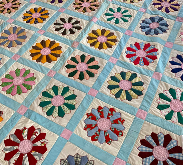 A colorful quilt with flower patterns sewn in each square, featuring various colors and designs, bordered with pastel blue and pink strips.