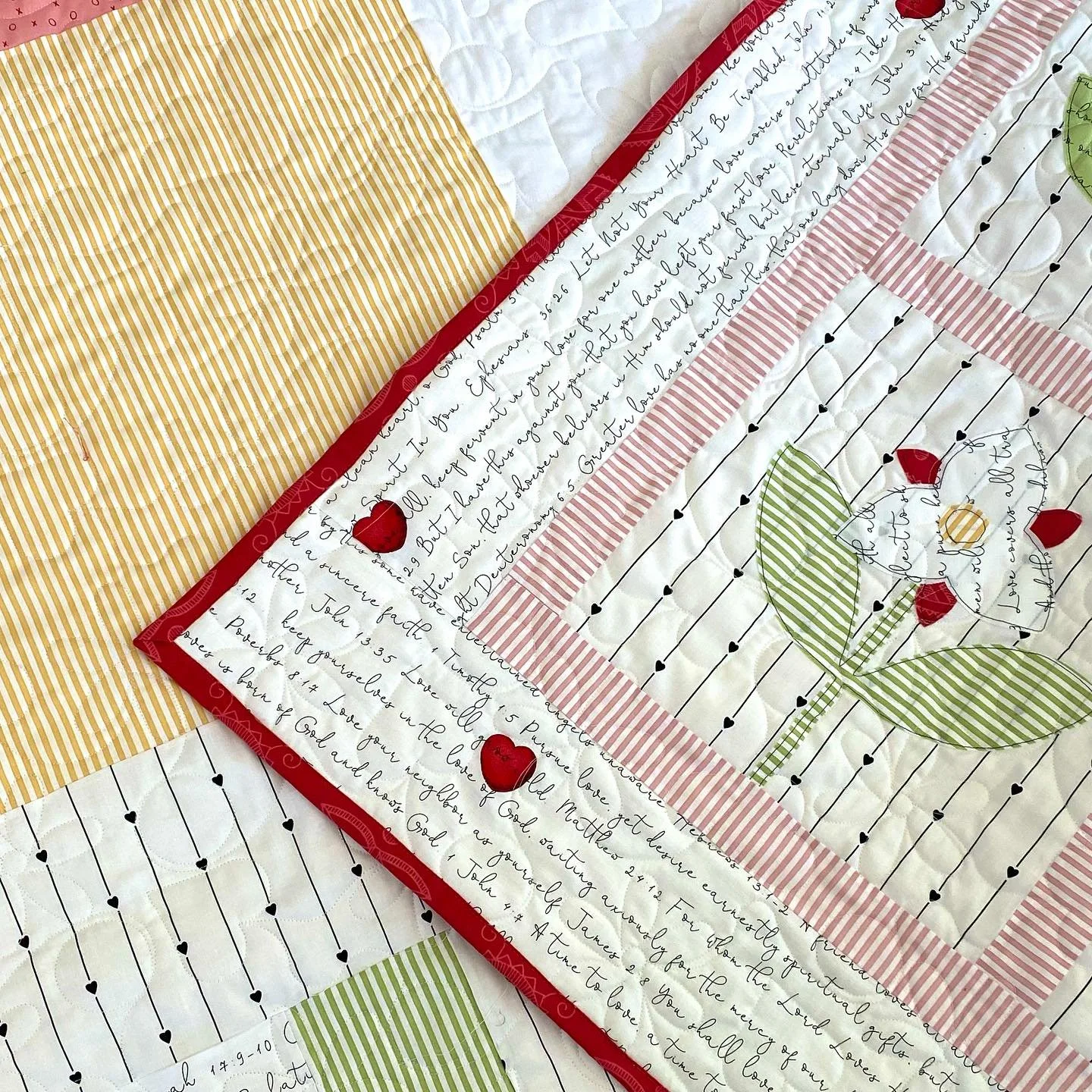 Close-up of a quilt with prayer phrases, embroidered hearts, and a flower design. The quilt includes various patterned fabrics in red, green, yellow, and white, with black details and red hearts.