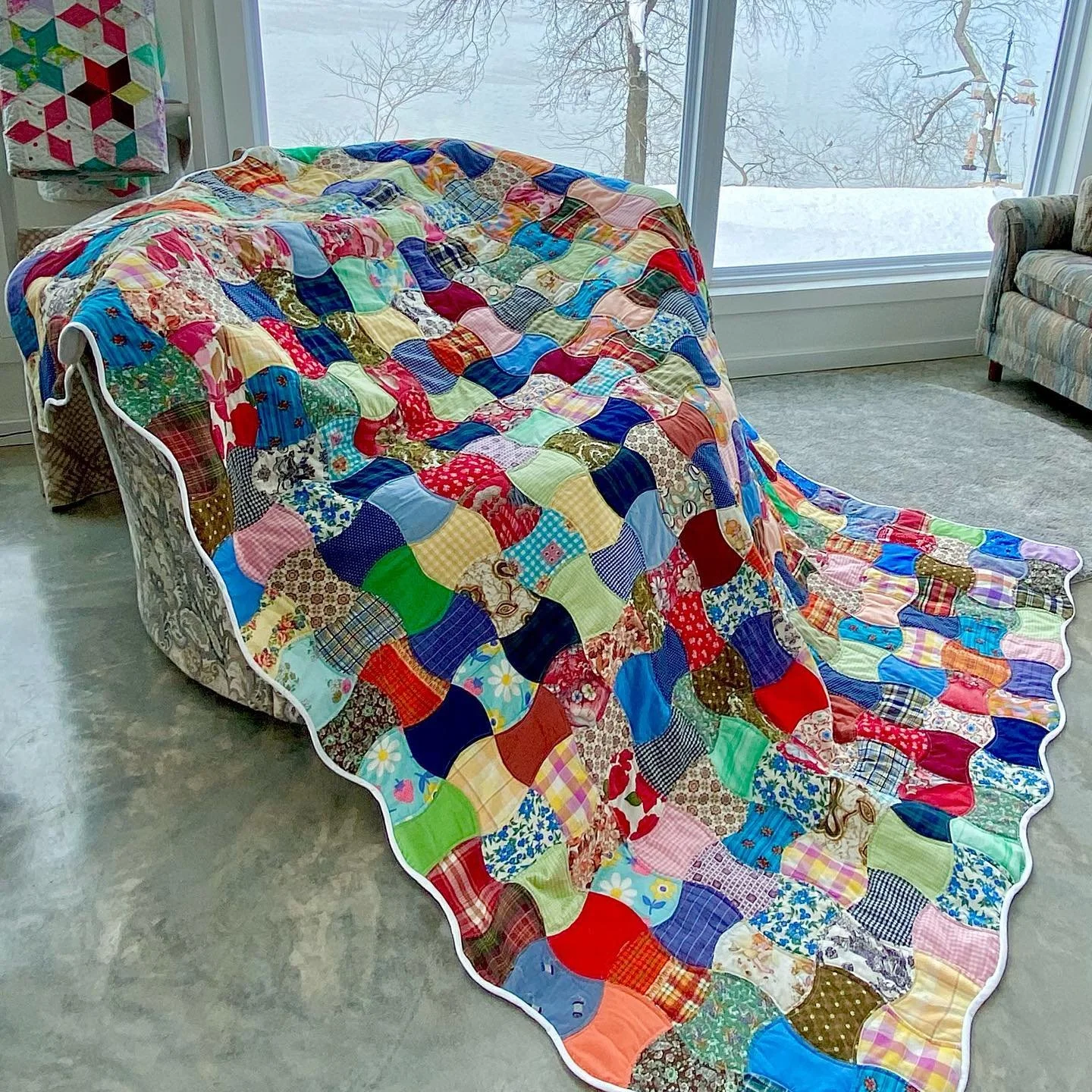 A colorful patchwork quilt draped over a sofa and flowing onto the floor, with a large window showing a snowy landscape outside.
