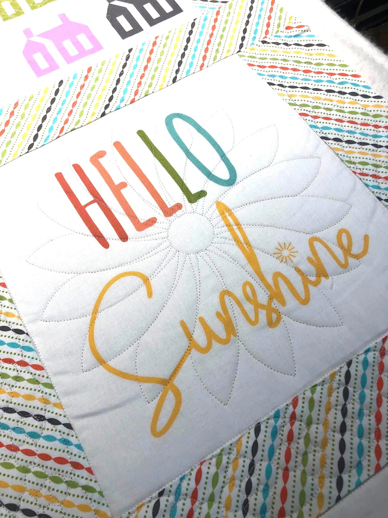 A colorful quilted fabric with the words 'Hello Sunshine' embroidered in bright, cheerful colors, featuring a large sunflower design.