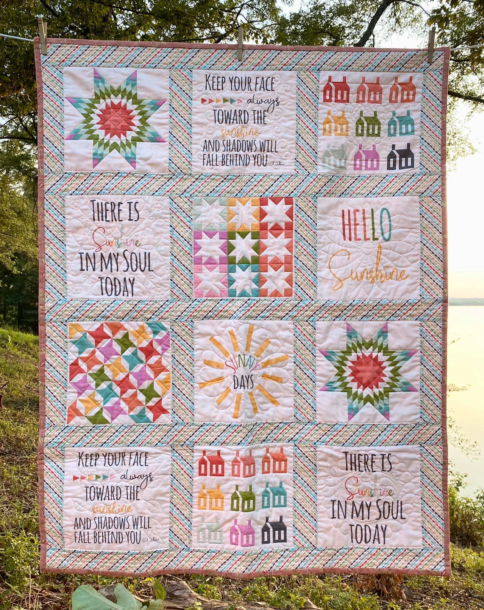 Colorful quilt hanging outdoors, featuring various quilt blocks with geometric patterns and inspirational quotes about sunshine and positivity.