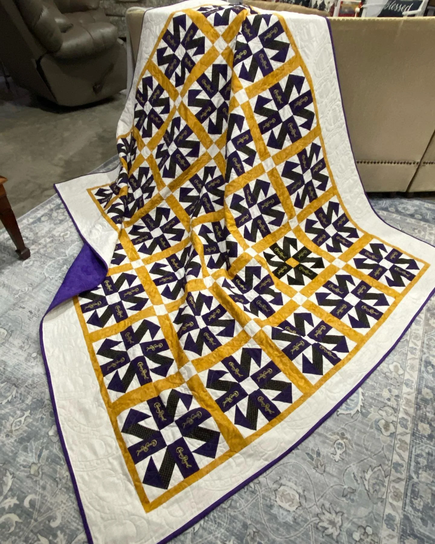 A decorative quilt with a purple, black, white, and gold pattern, featuring the word 'Gator' and a key design, draped over furniture in a living room.