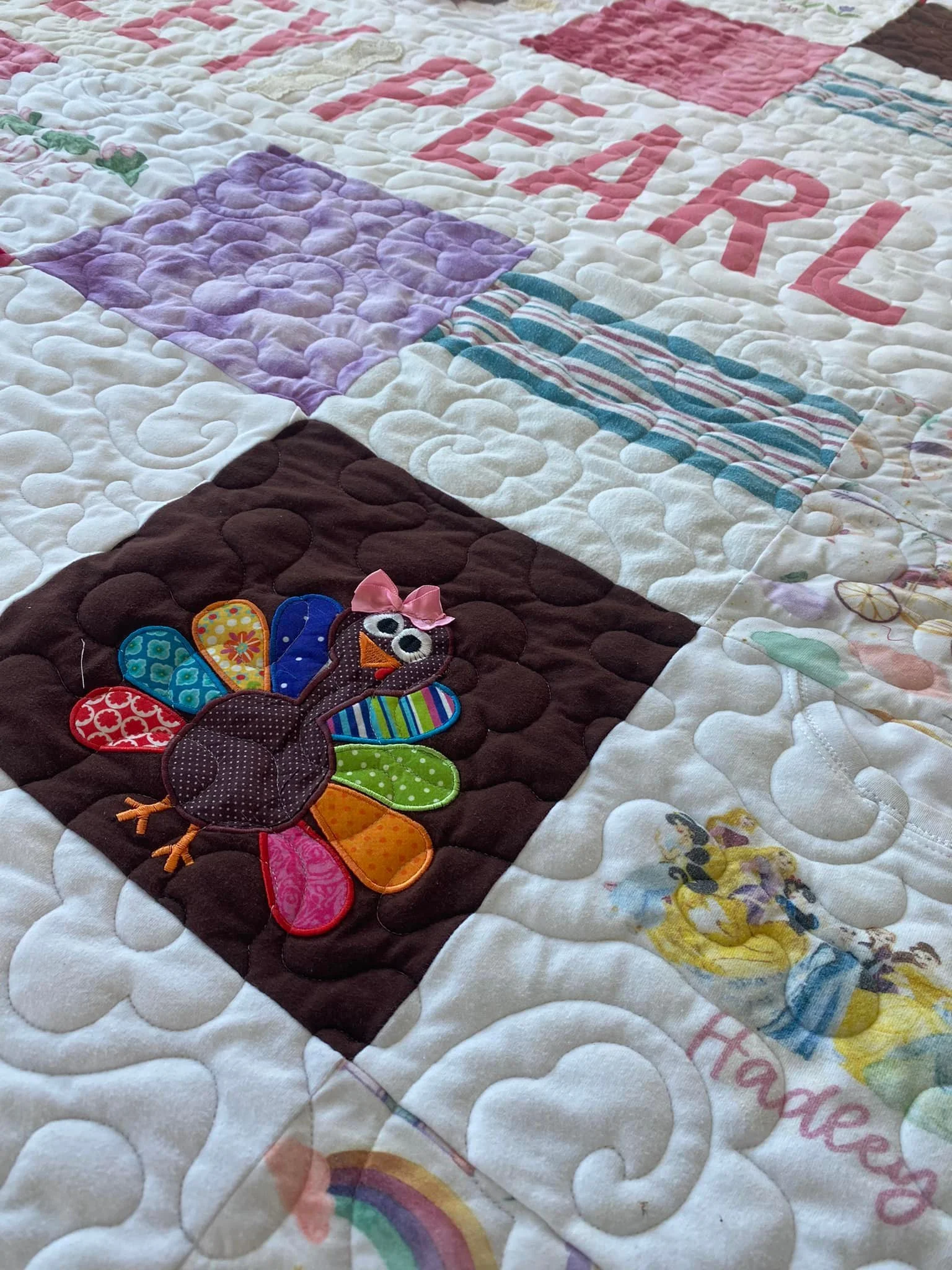 Close-up of a colorful quilted blanket with various patterns, including a turkey appliqué with rainbow-colored feathers and a pink bow, and parts of Disney princess images.