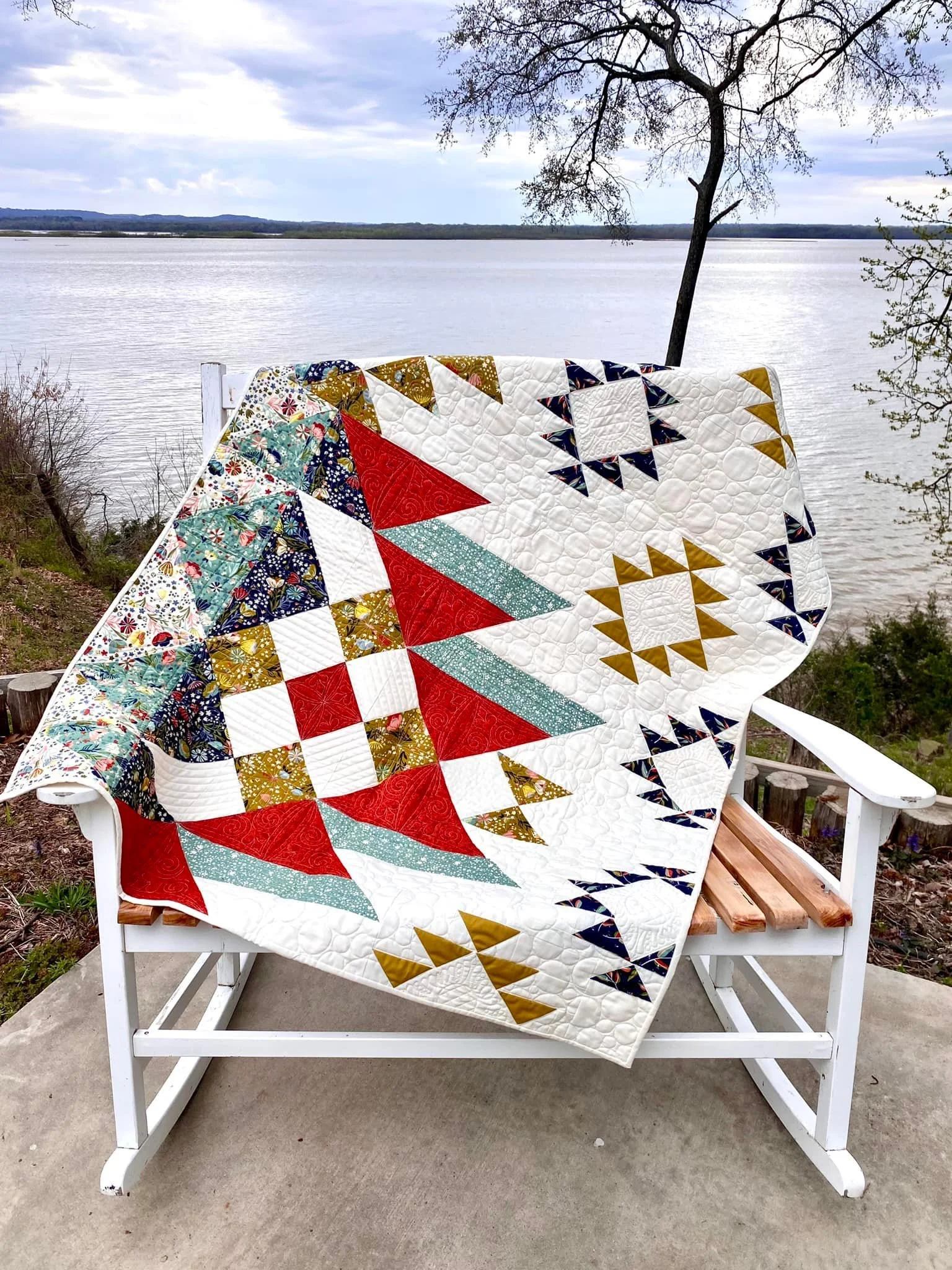 A colorful quilt with geometric patterns draped over a white wooden rocking chair near a river, with trees and a cloudy sky in the background.